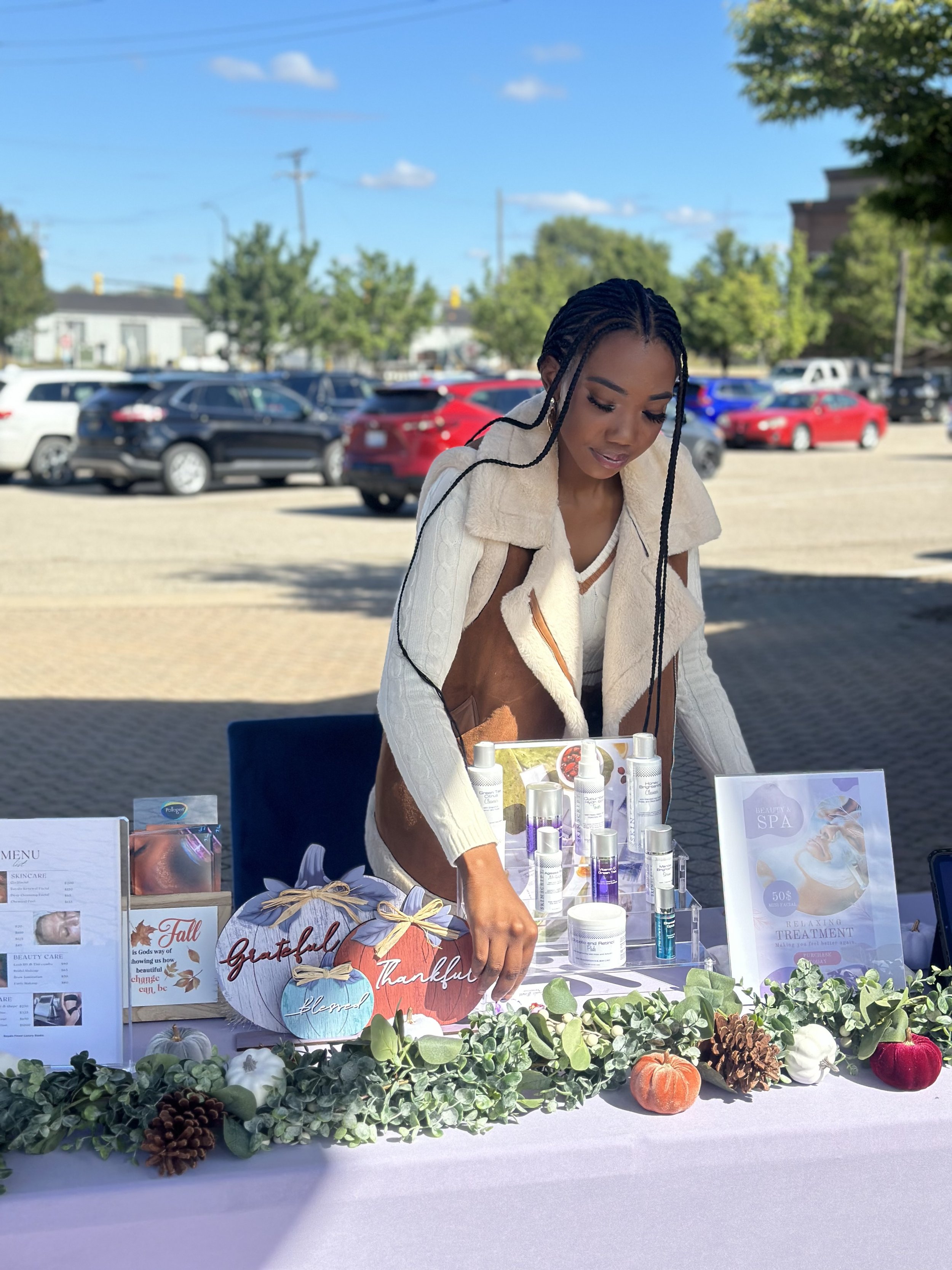 A woman with braided hair stands behind a table covered with skincare products. The table is adorned with decorative pumpkins and greenery. A spa treatment menu and product flyers are also displayed. Several cars are parked in the background on a sunny day.