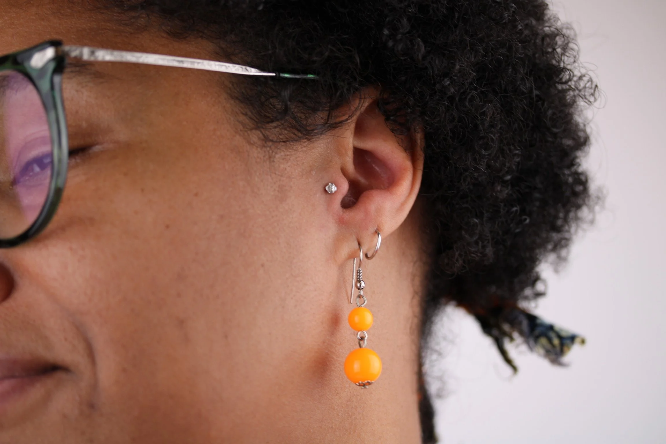 Close-up of a person's ear with multiple earrings, including a diamond stud, a small hoop, and a dangle earring with two orange beads.