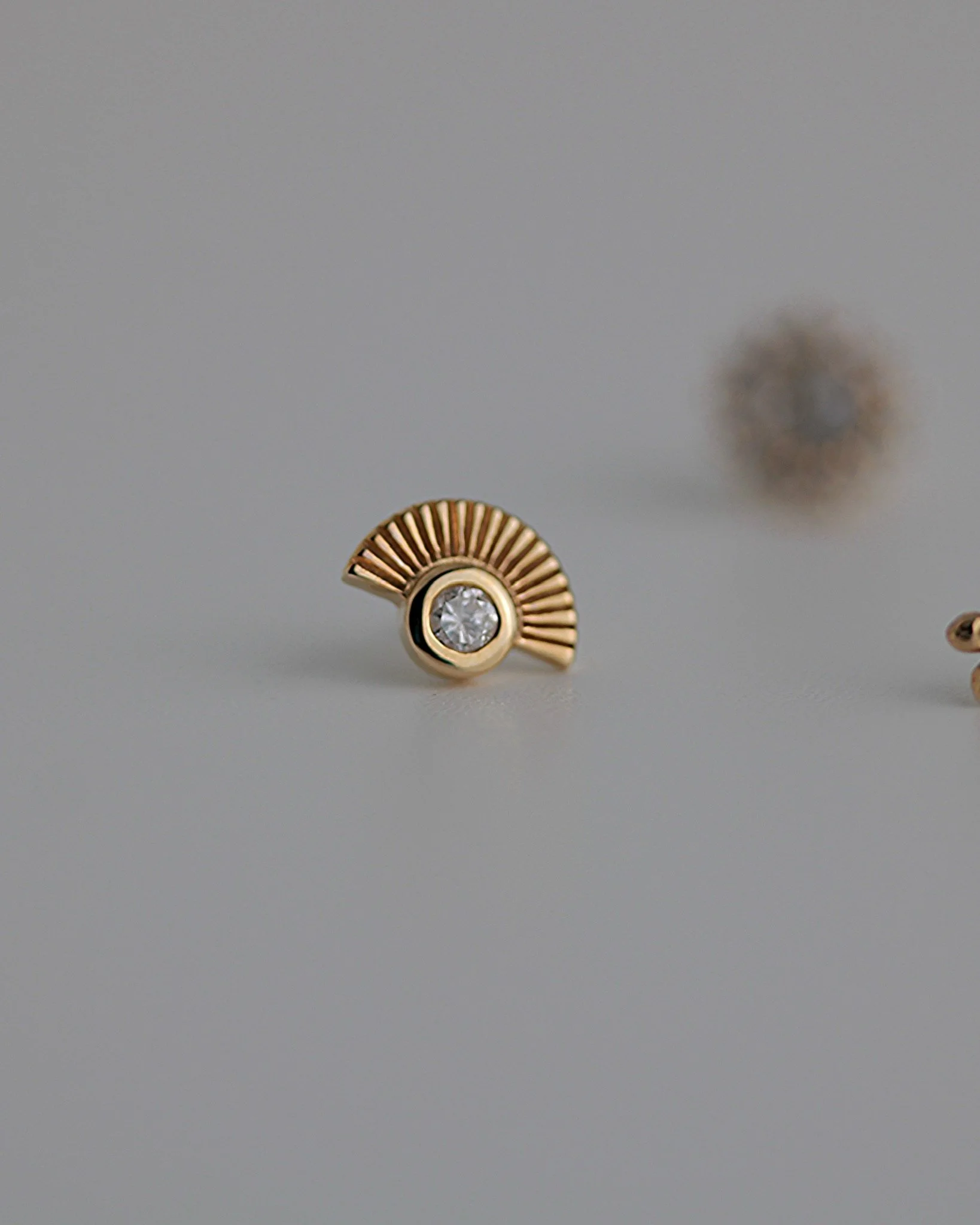 A gold earring with a circular diamond in the center and a fan-shaped gold design composed of thin, radiating bars.