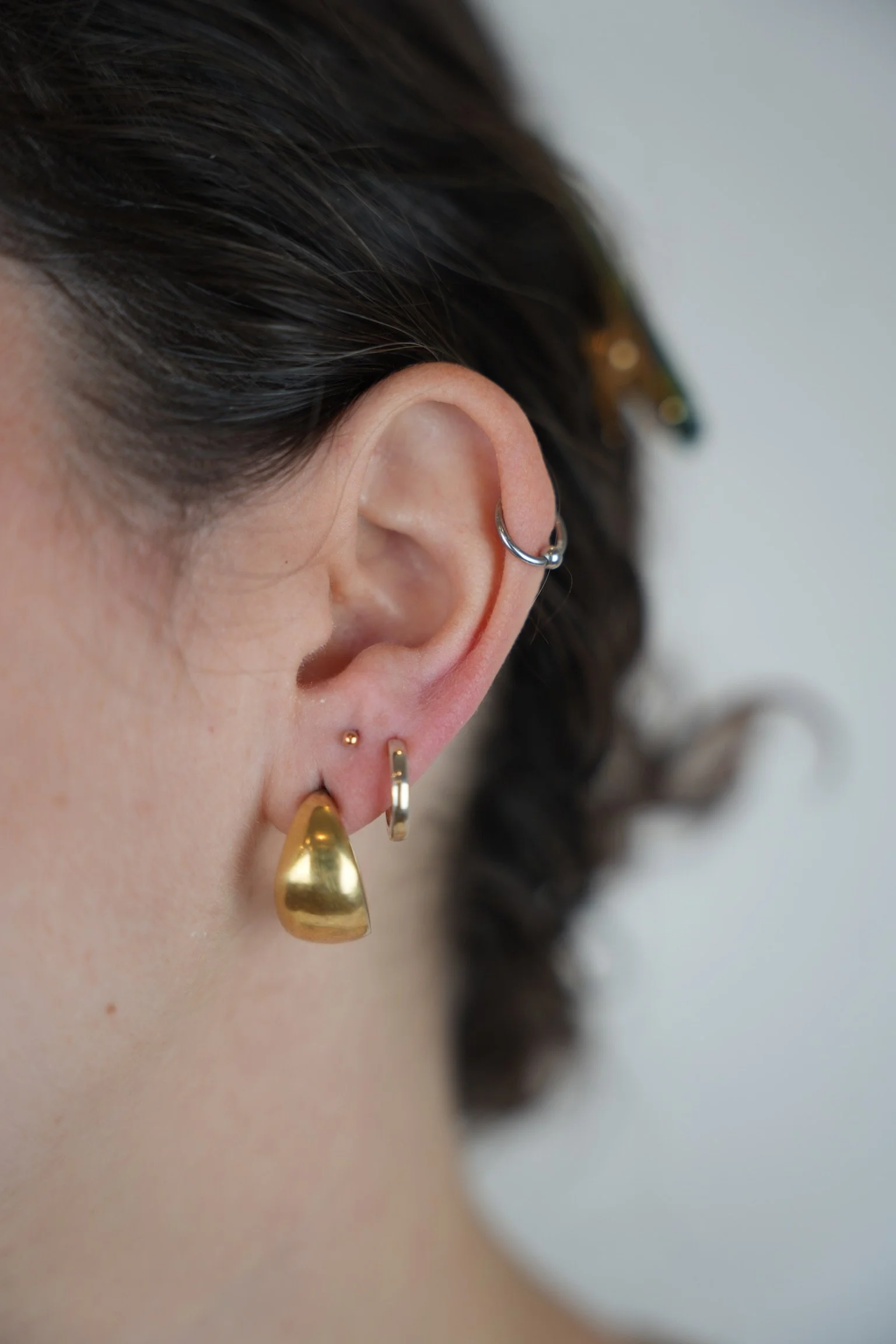 Close-up of a person's ear with multiple earrings, including gold, silver, and small stud earrings, and dark brown hair styled with waves.