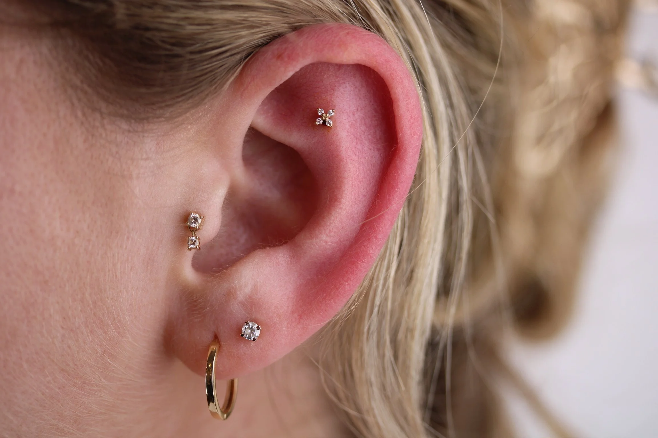 Close-up of a person's ear with multiple piercings, including a gold hoop in the earlobe and three additional earrings with gemstones in the upper and middle ear.