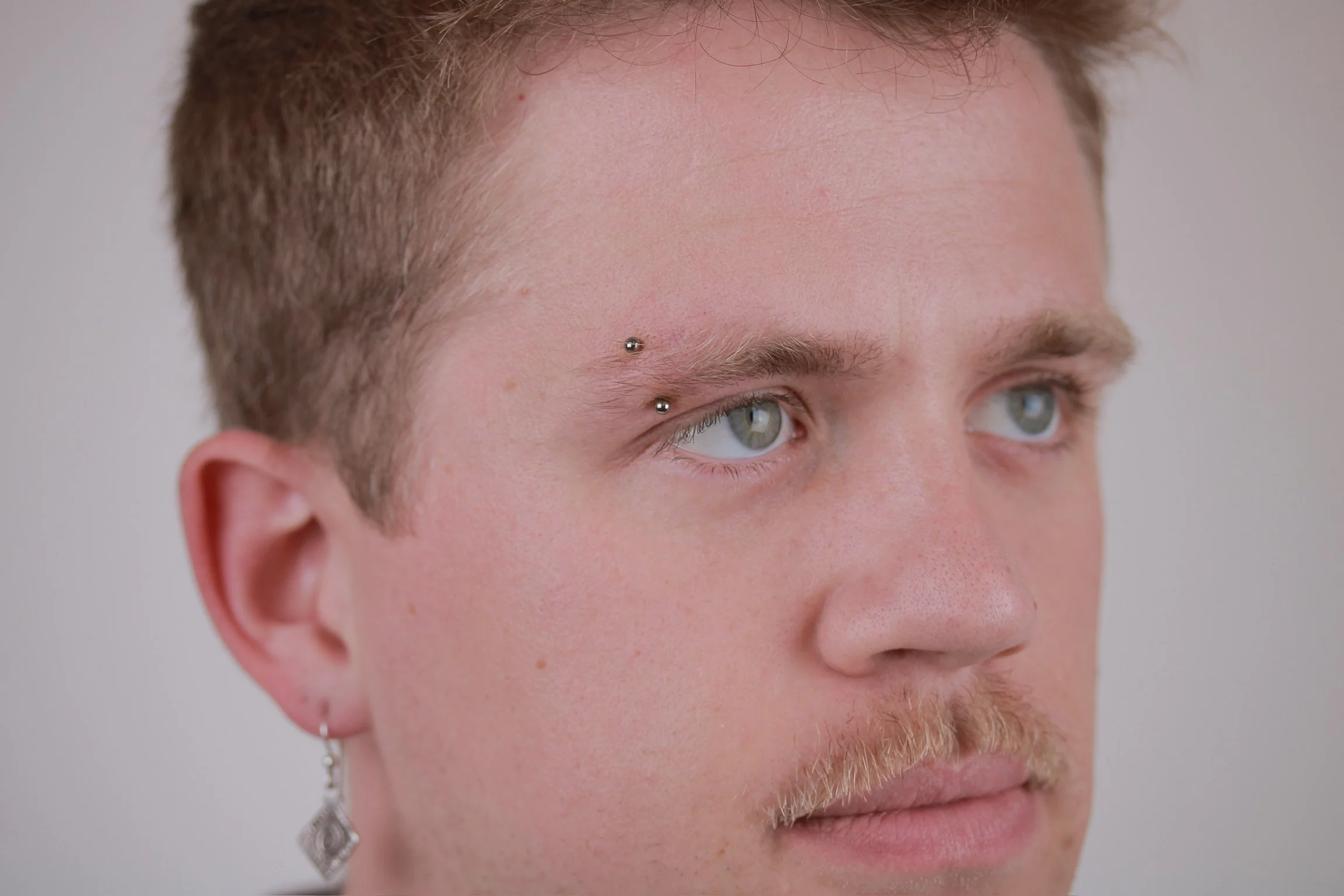 Close-up of a young man with short, light brown hair, piercing blue eyes, and a light beard, featuring two silver dermal piercings above his right eyebrow and an earring in his left ear.