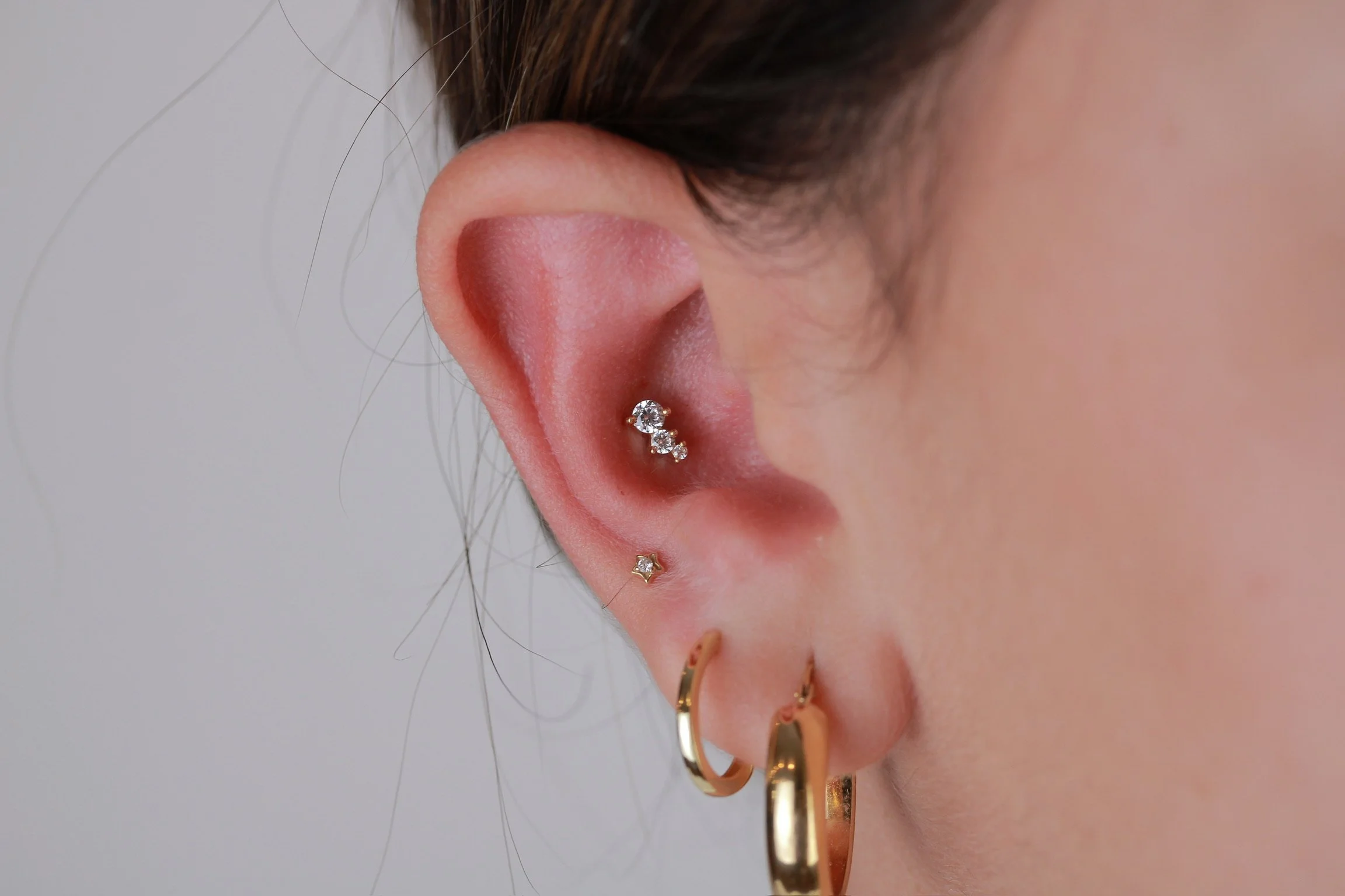 Close-up of a person's ear with multiple earrings, including a small gold hoop, a small gold stud, and two silver earrings with clear gemstones.