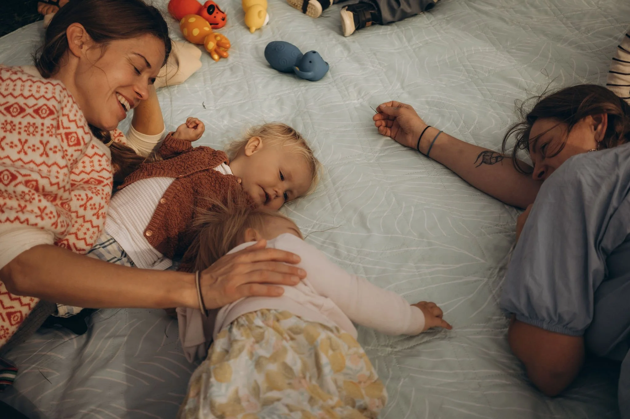 Two women and two children lying on a bed, smiling and interacting with each other. The children are in the middle, one leaning on the mother on the left, and the other lying on her stomach. The women are on each side of the children, one on the left and the other on the right. There are colorful toys on the bed.