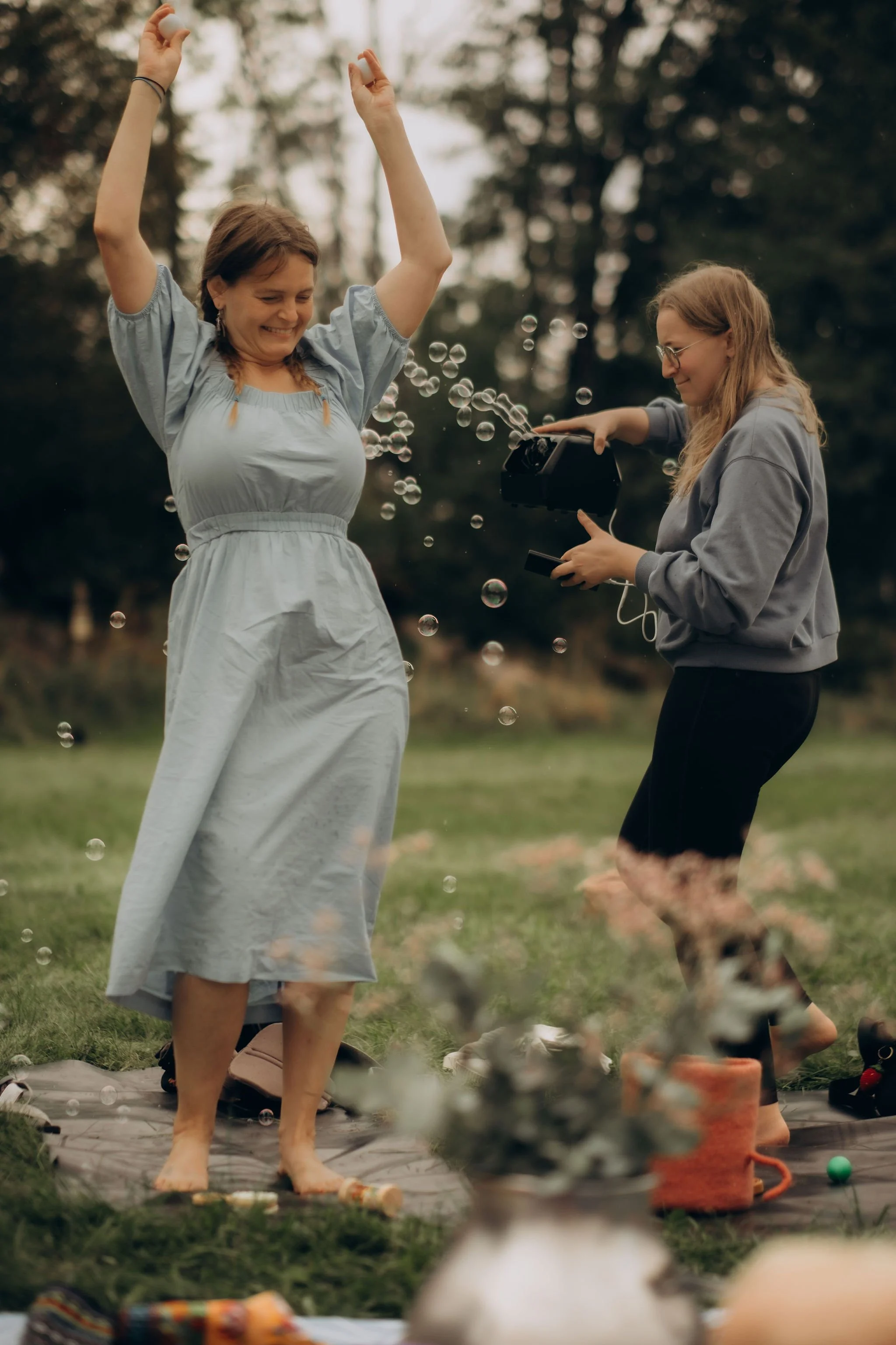Two women are enjoying a picnic outdoors; one is standing with arms raised, smiling, while the other is taking a photo. There are bubbles floating around, and a blanket with various picnic items like drinks and flowers on the grass.