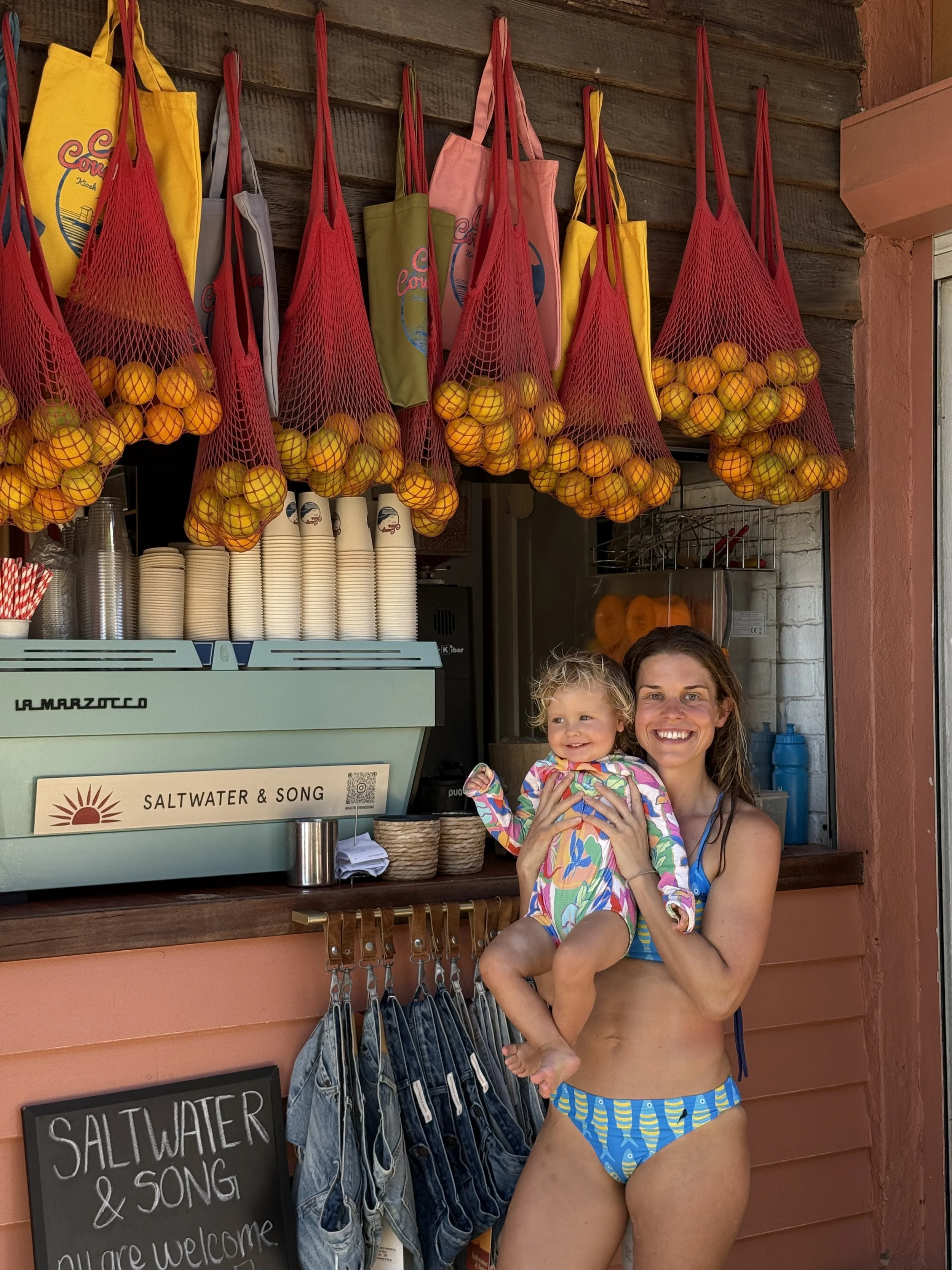 Woman in a bikini holding a smiling little girl in a colorful swimsuit in front of a fruit stand with hanging bags of oranges, a coffee machine, paper cups, and jeans on display.