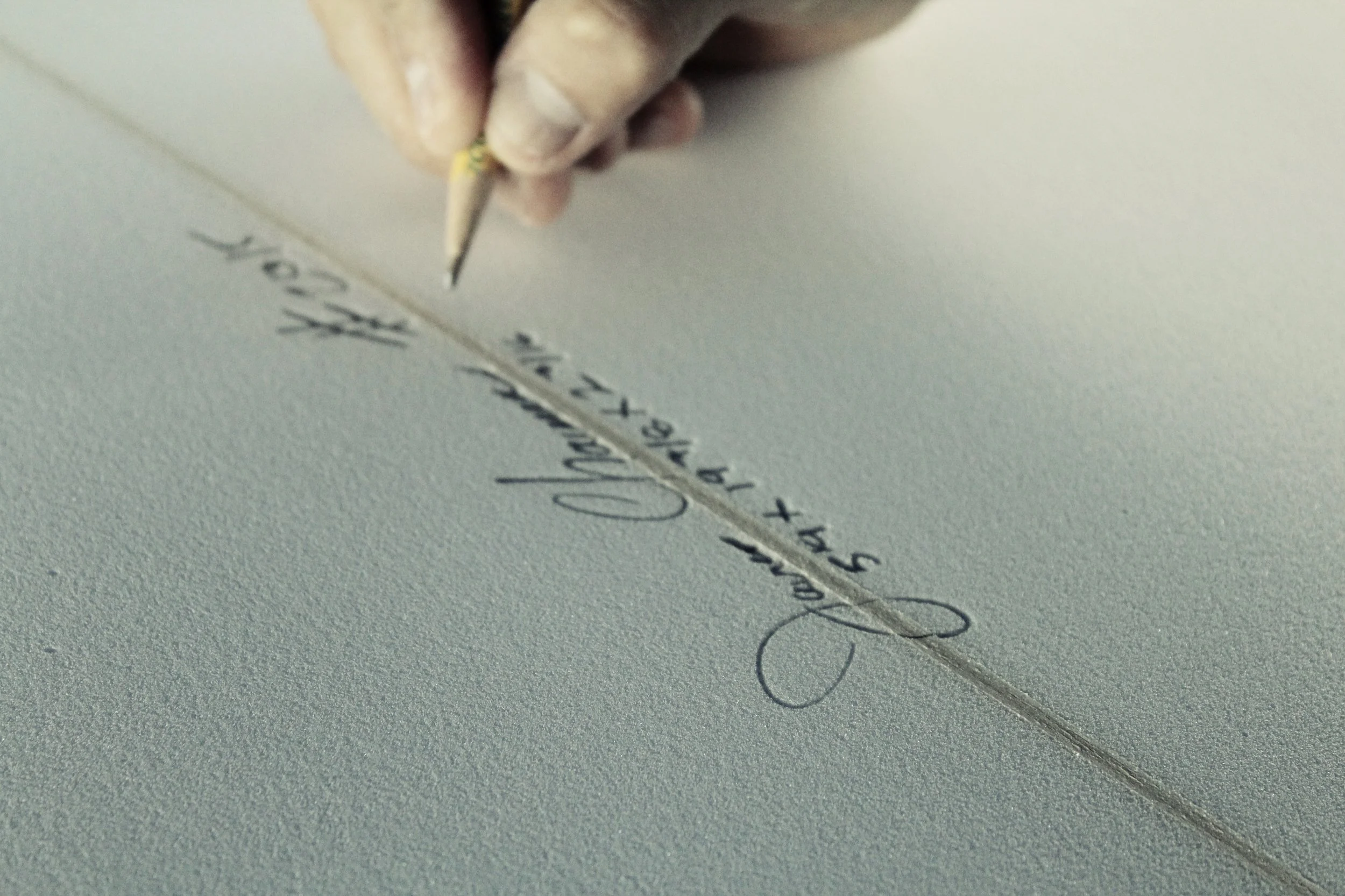 Primer plano de una mano que sostiene un lápiz, escribiendo texto cursivo en una tabla de surf.