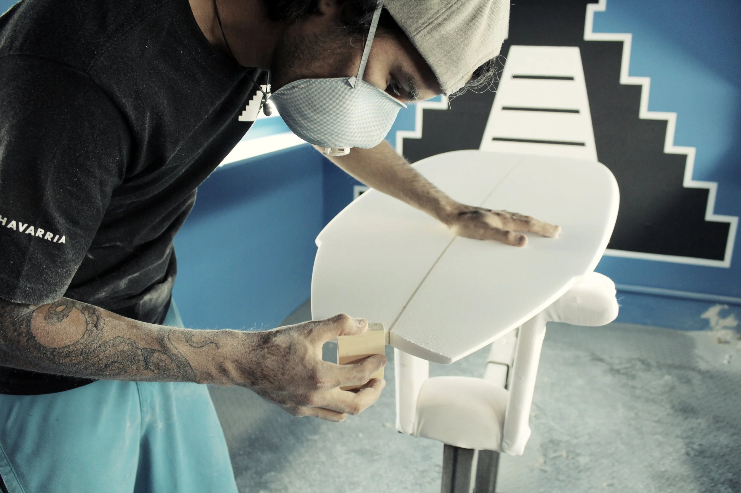 Un hombre trabajando en la conformación de una tabla de surf, con una máscara y gafas, en un taller con una pared azul y una obra de arte estilizada de una pirámide escalonada en blanco y negro: el logotipo de Mainland Surfboards