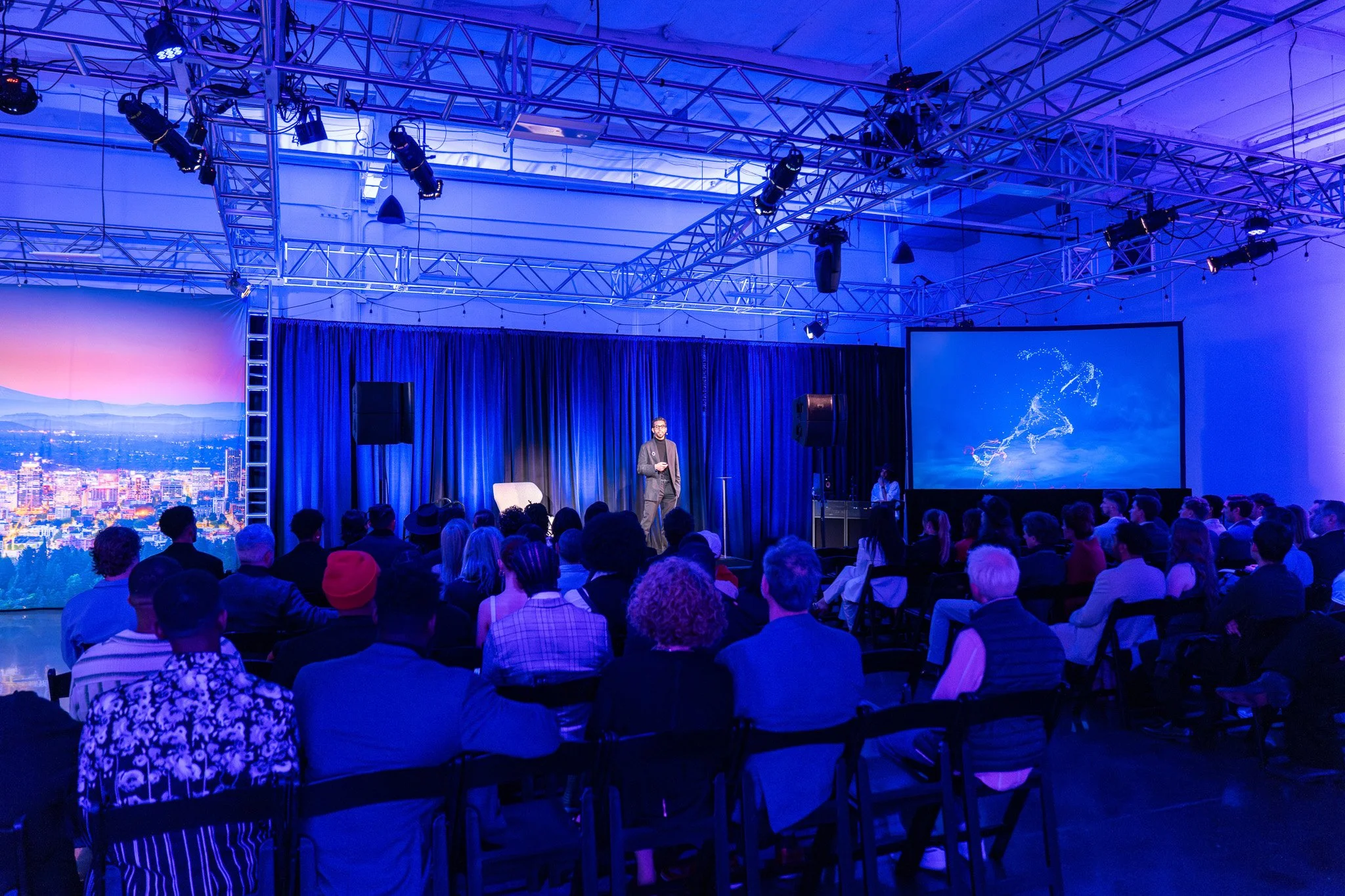 A speaker on stage at a conference or event, addressing an audience seated in front. The stage has black curtains and is illuminated with blue lighting, and a large screen displaying visuals is to the right of the speaker. The audience is diverse, facing the stage attentively.