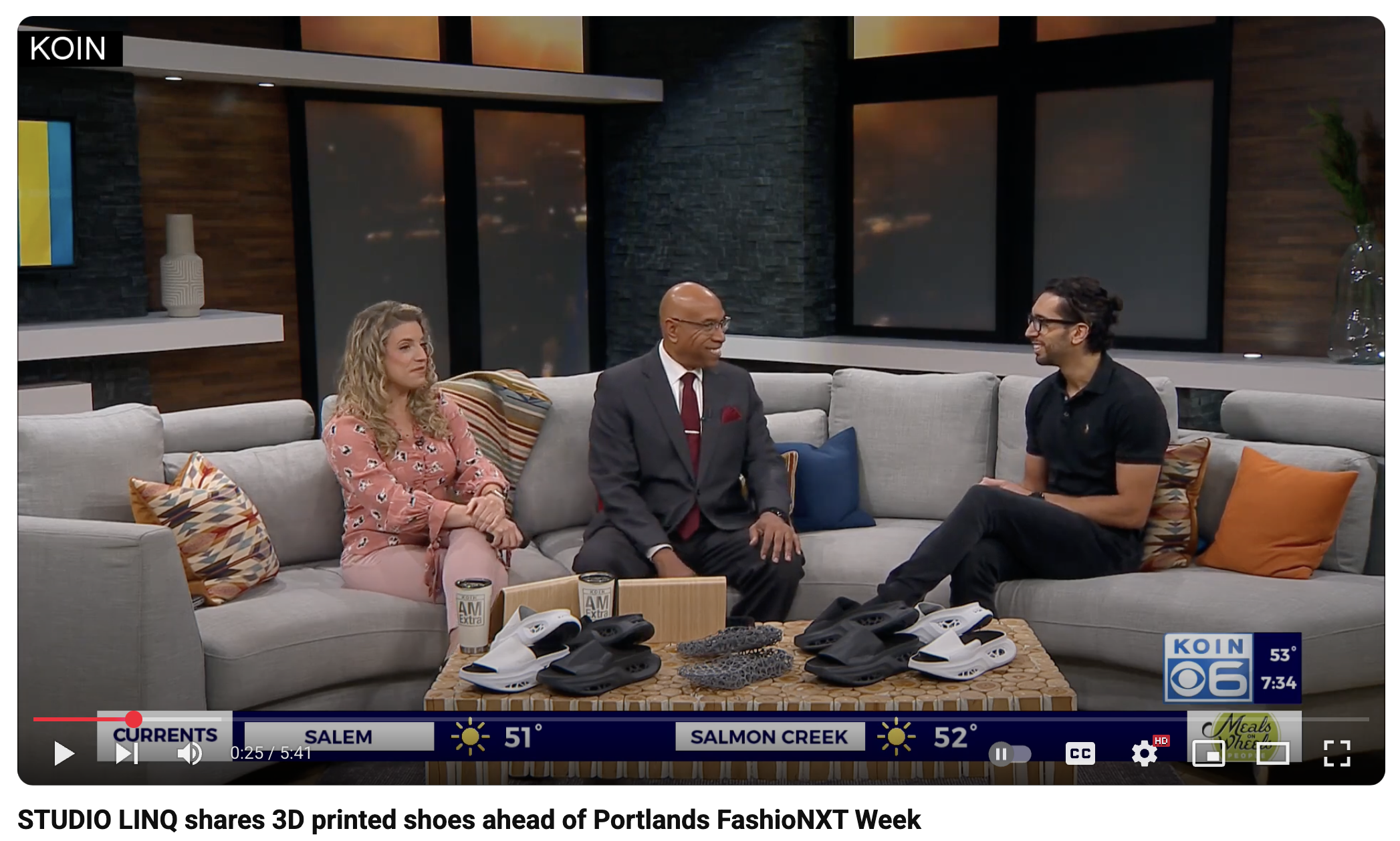 Three people sitting on a light gray sectional sofa in a television studio with dark wood and stone walls, colorful pillows, and a wooden coffee table with 3D printed shoes and coffee mugs in front of them, during a news segment.