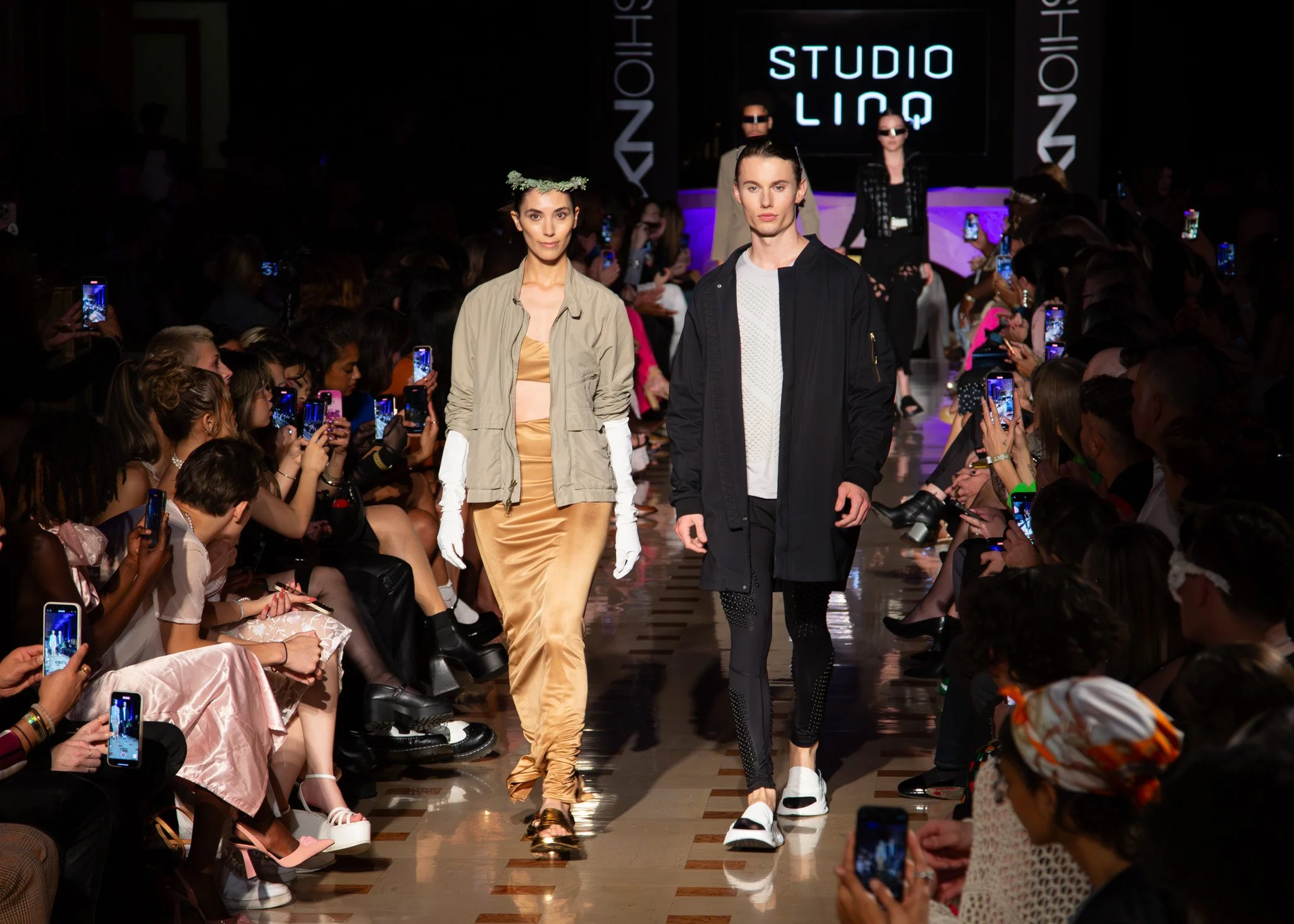 Models walking down the runway at a fashion show, with an audience taking photos. The background features a sign that says "STUDIO LINK."
