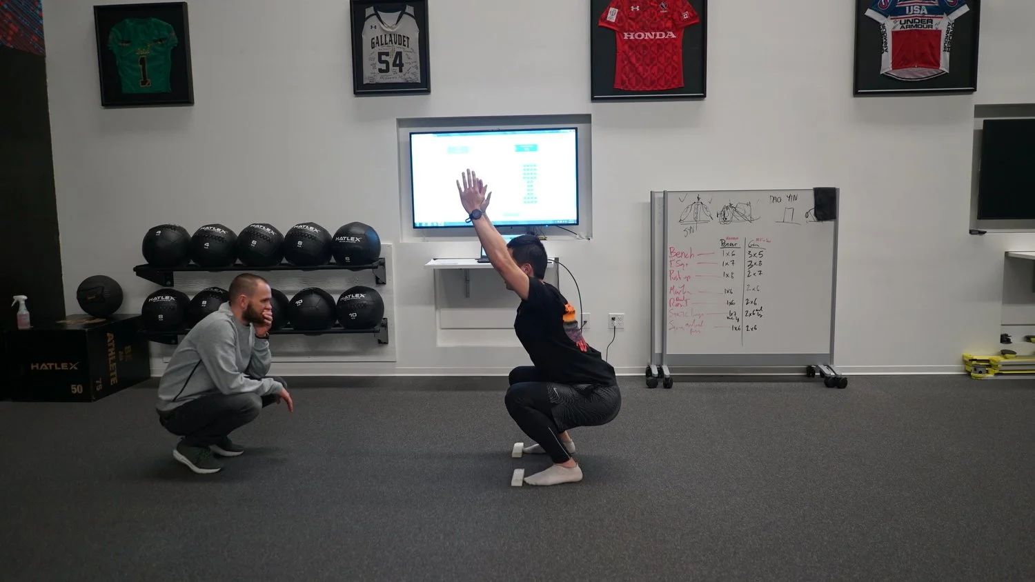 A woman doing squats in an indoor gym with a trainer kneeling and watching her. There are framed sports jerseys on the wall, a row of medicine balls, a whiteboard with workout notes, and a monitor displaying a hand gesture.