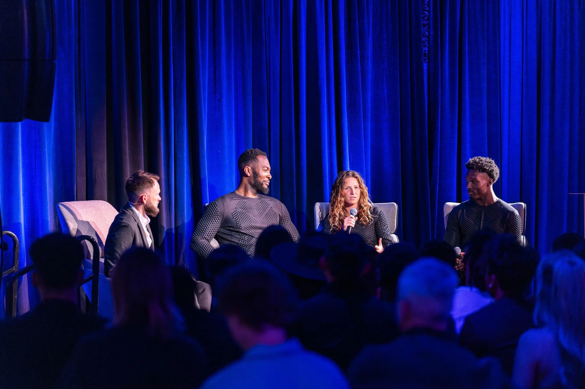 Four people on a stage, one woman using a microphone, against a blue curtain backdrop, with an audience in front.