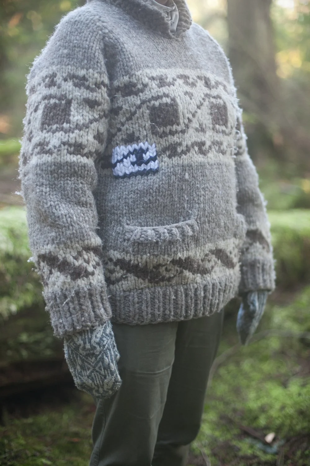 A person wears a cowichan sweater while standing in the forest. The cowichan has a visible mend on the right chest.