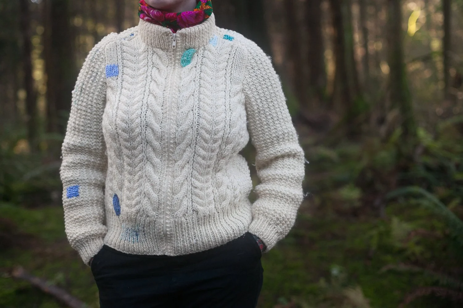 a white cable sweater with blue, purple, and green darns.