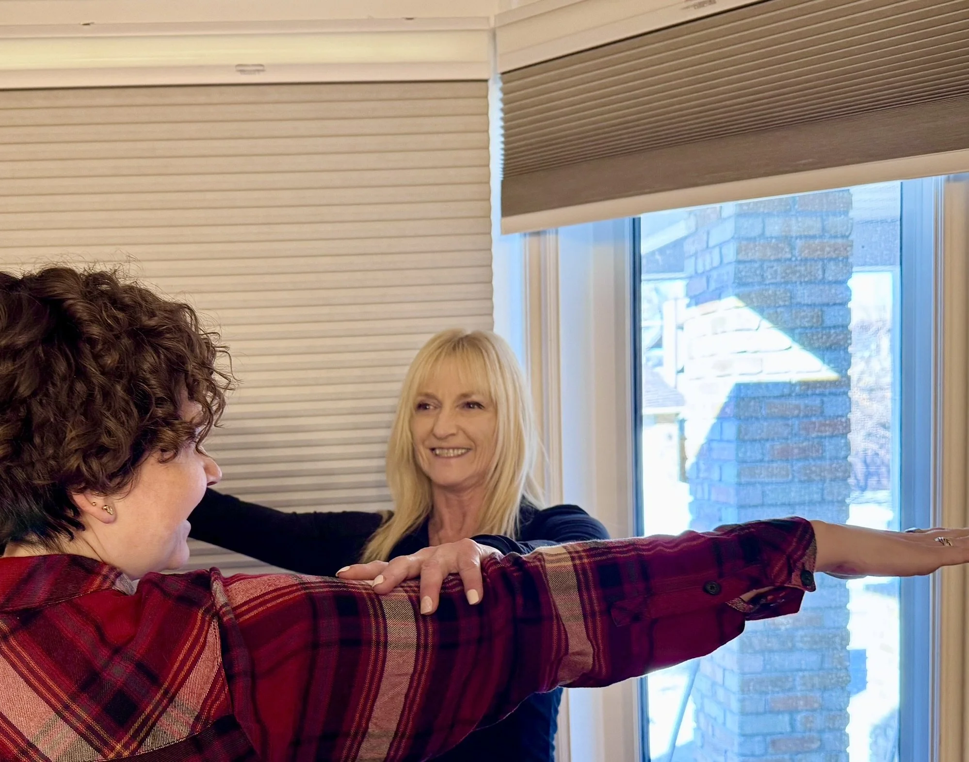 Two elderly women standing in a kitchen doing exercise, increasing flexibility and raising their arms, with a personal trainer at home. The kitchen has dark wood cabinets and stainless steel appliances.