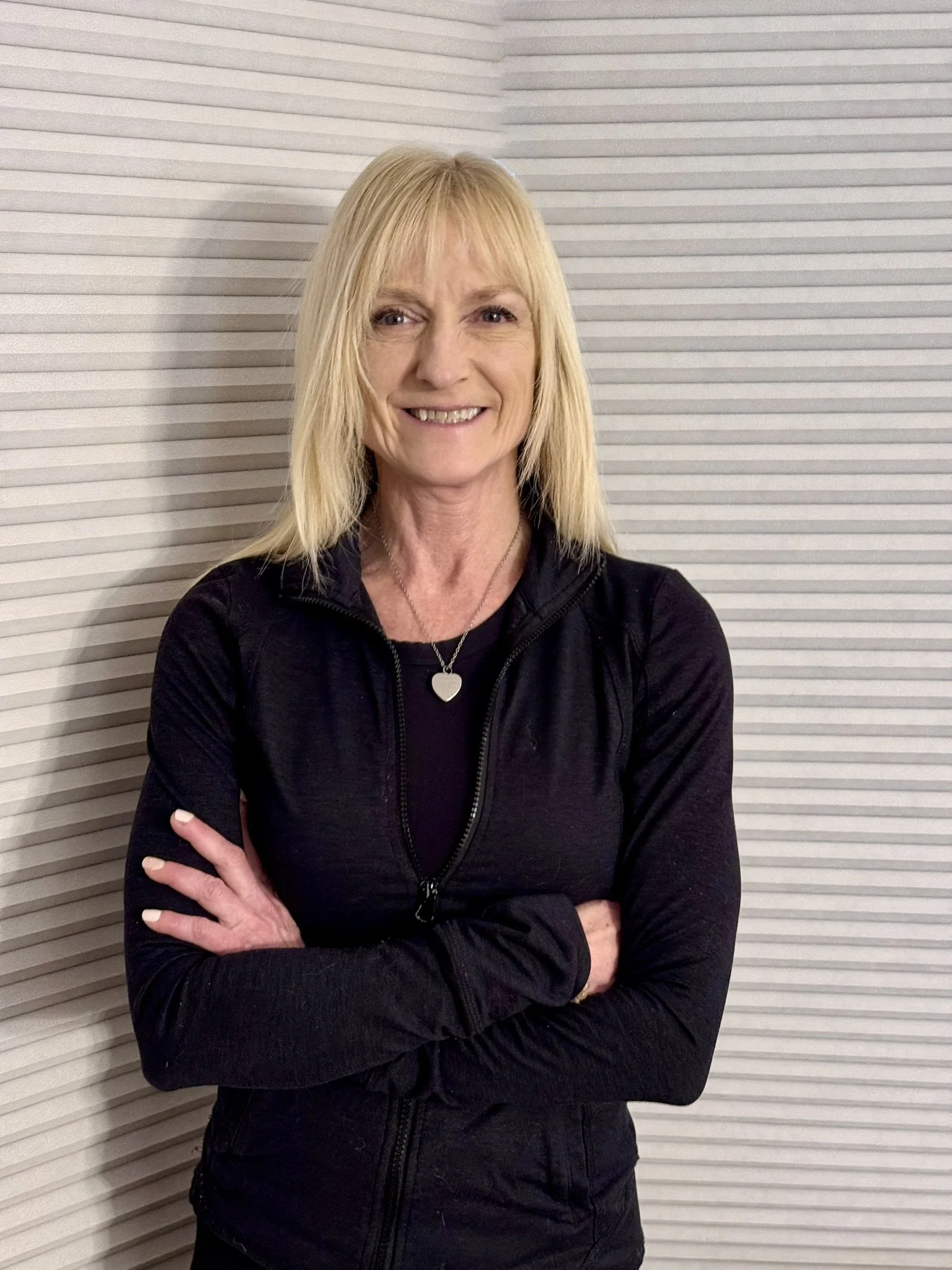 Smiling personal trainer with blonde hair and a black jacket standing against a striped background.