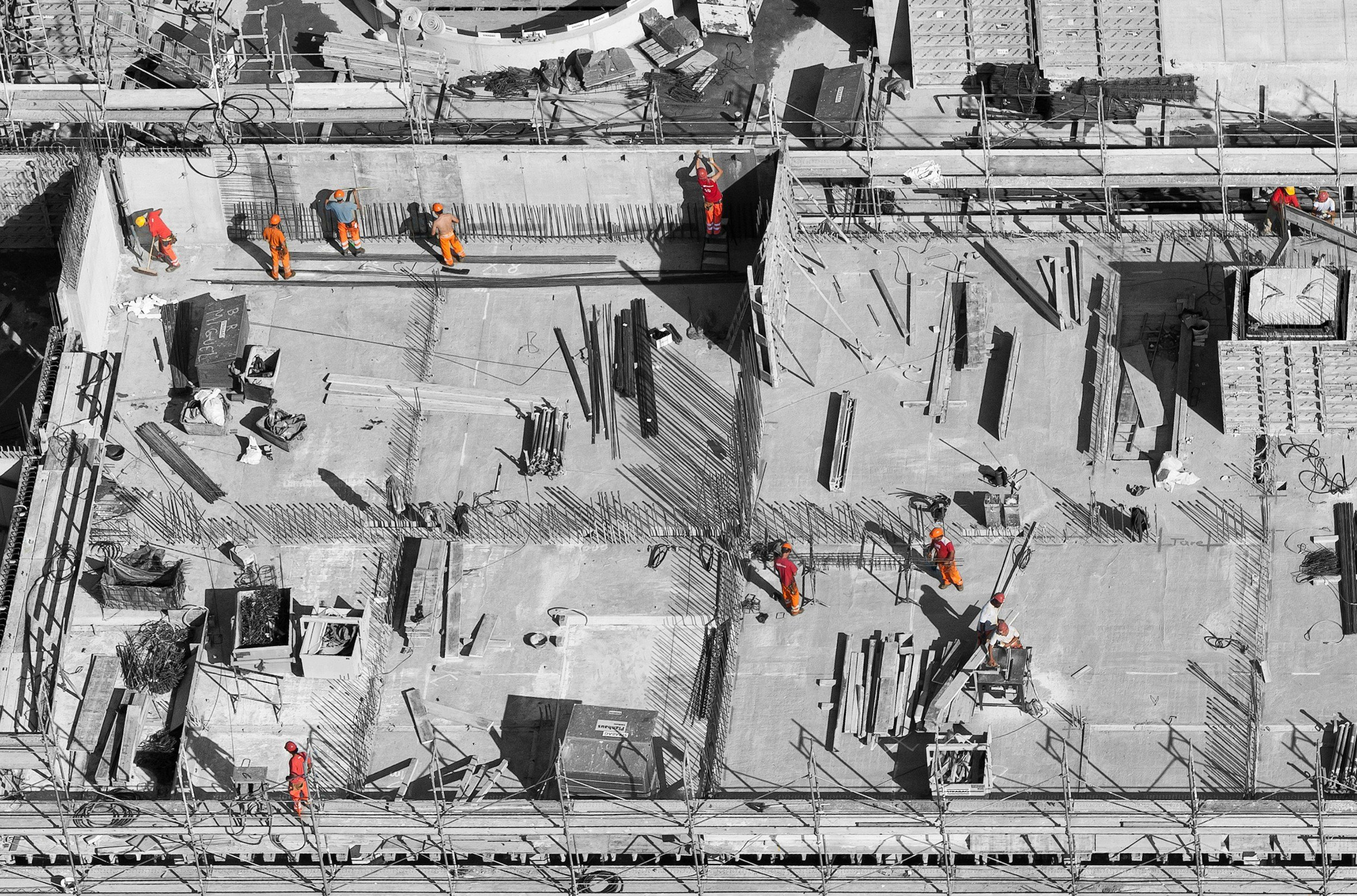 High-angle view of a construction site with workers in orange safety gear working on the concrete foundation and walls, surrounded by scaffolding and construction materials.