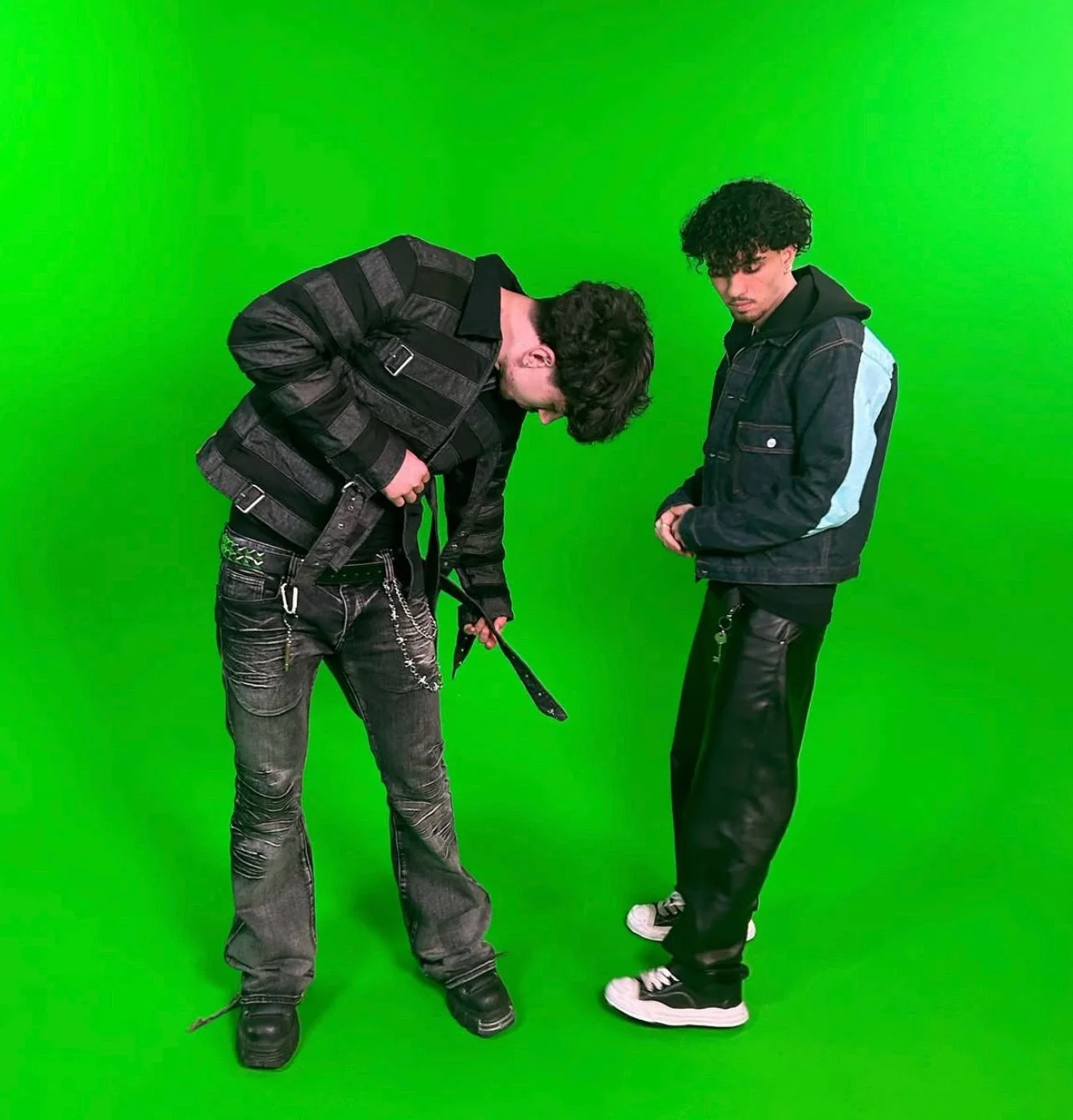 Two young men standing in front of a green screen, one looking down and adjusting his belt, the other looking at the ground with hands clasped.