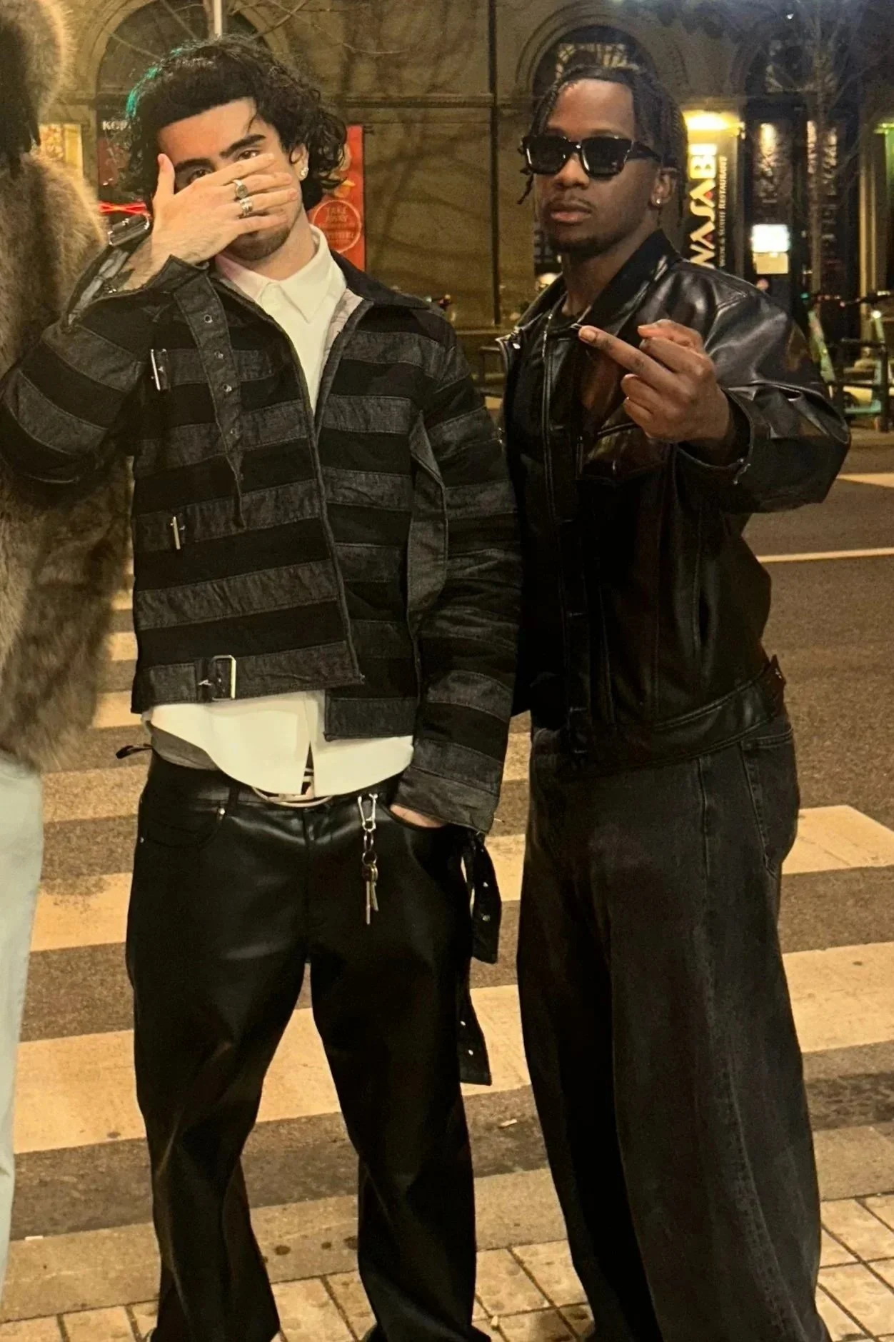 Two men standing on a city street at night, one covering his mouth with his hand and the other wearing sunglasses, both dressed in black leather jackets.