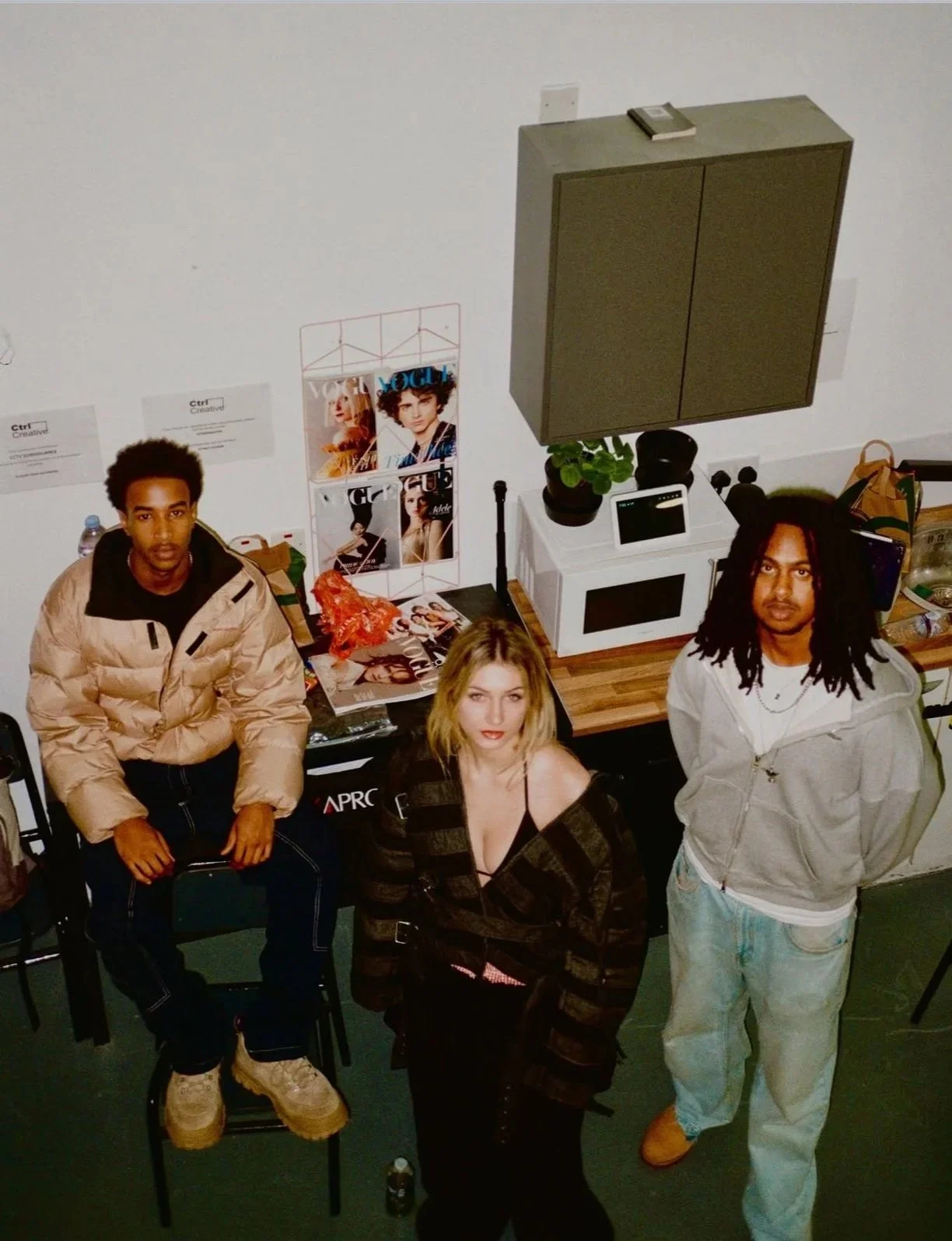 Three young people standing in a room with a kitchen area and magazines, with a high angle view of the group.