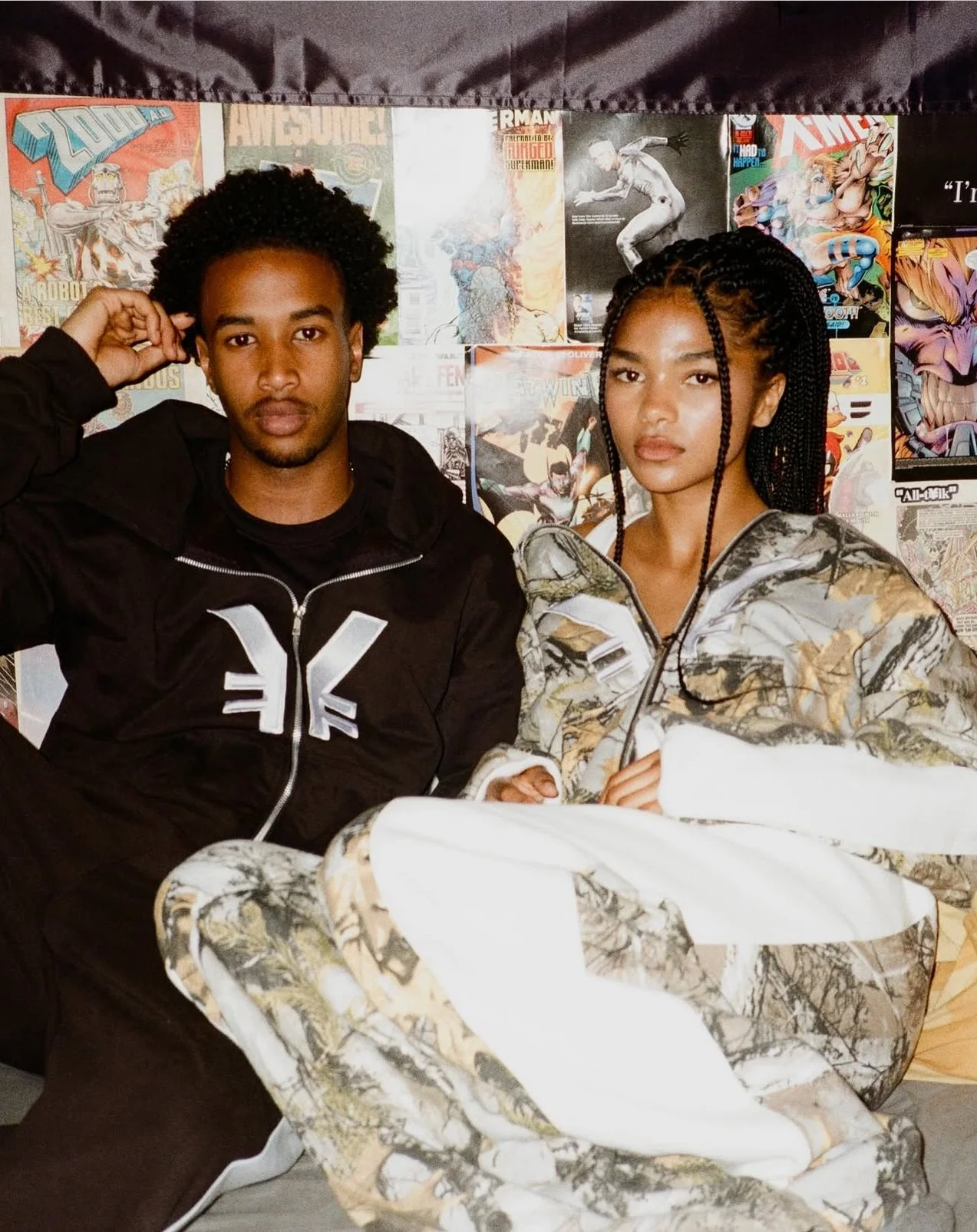 Two young adults, a man and a woman, sitting against a collage of comic book covers and posters background. The man has curly hair, wearing a black hoodie with a white logo, and is touching his head. The woman has long braided hair, wears a camouflag
