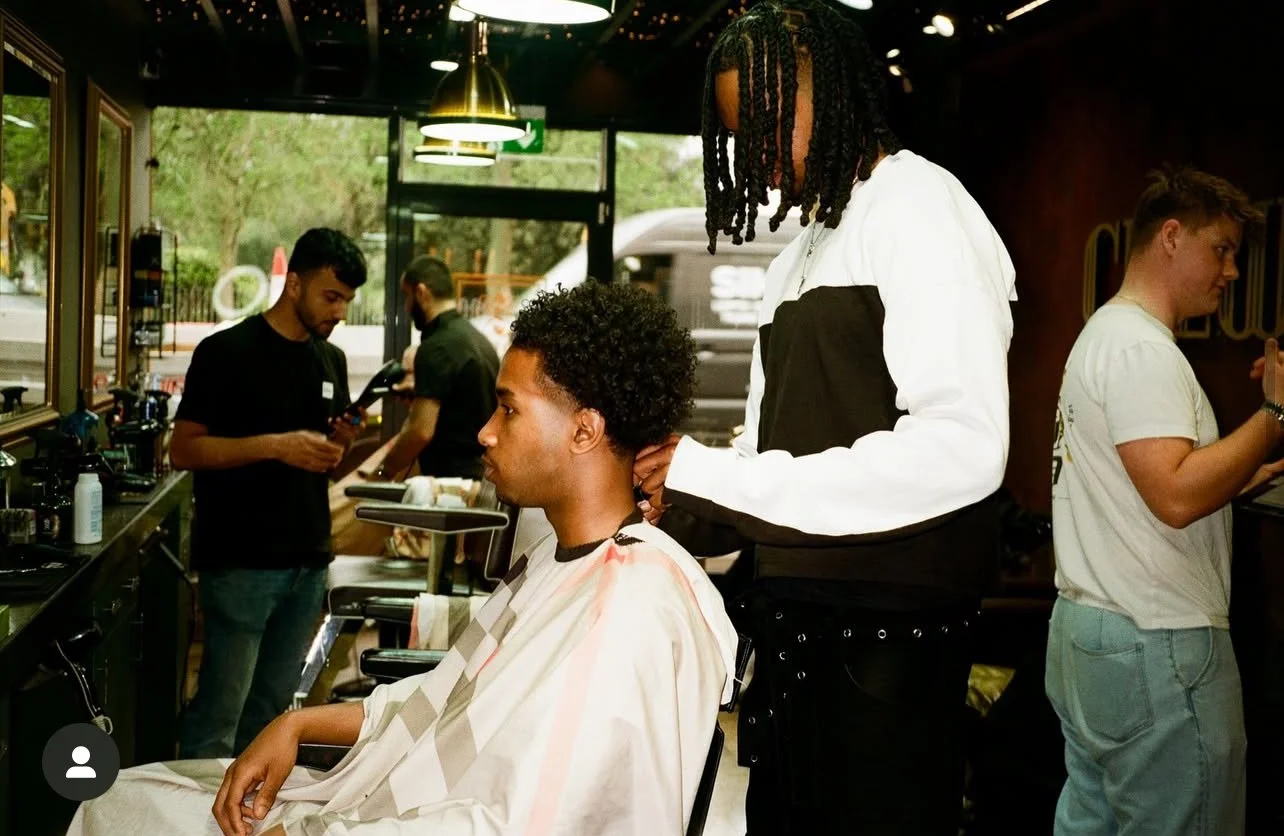 A man with curly hair getting a haircut in a barbershop while another man with dreadlocks stands behind him. Two other people are in the background using their phones.