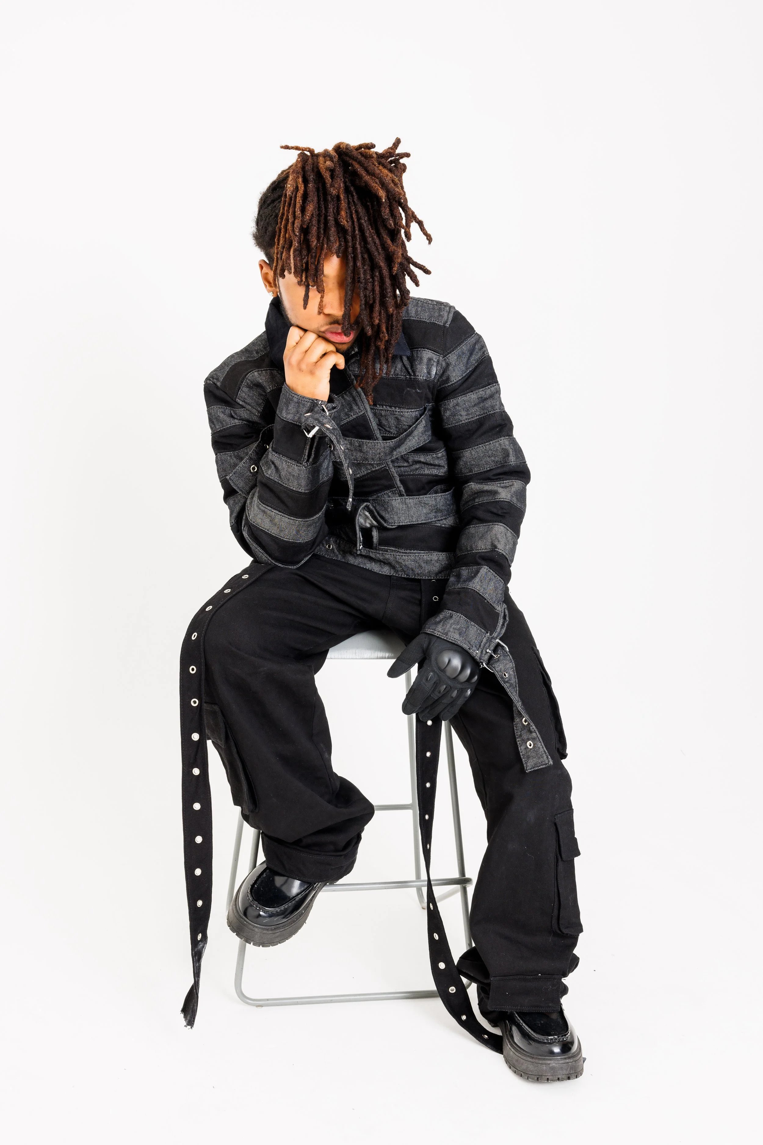 Young person with dreadlocks sitting on a stool, wearing black and gray striped jacket, black pants with silver grommets, black gloves and shiny black boots, against a plain white background.