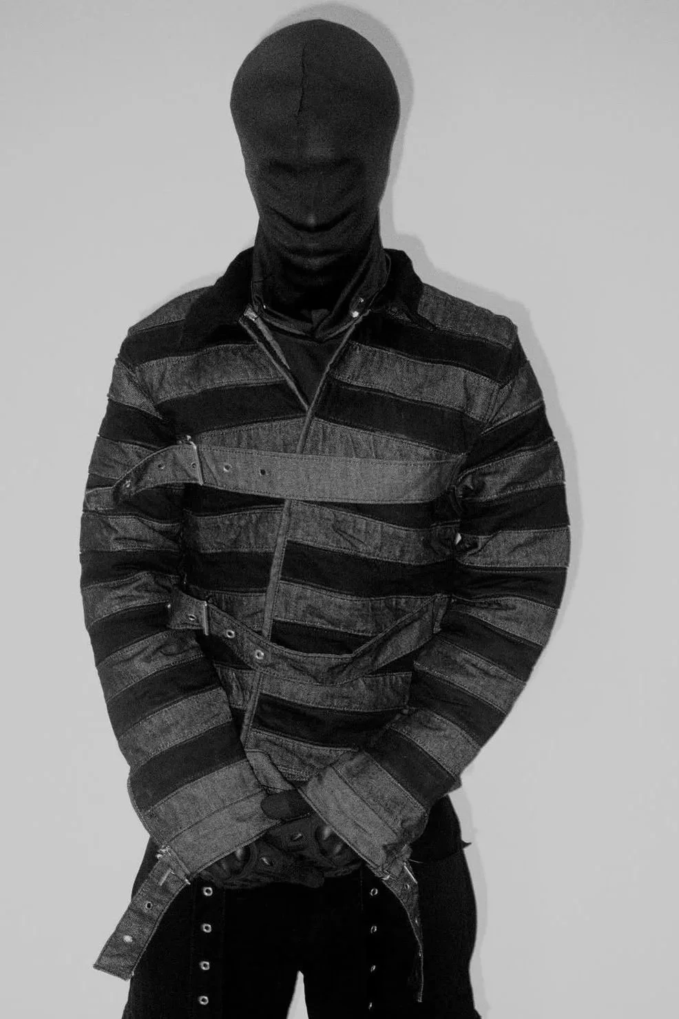 A person wearing a black ski mask, a striped jacket with belt details, and black pants with metal eyelets, standing against a plain white background.
