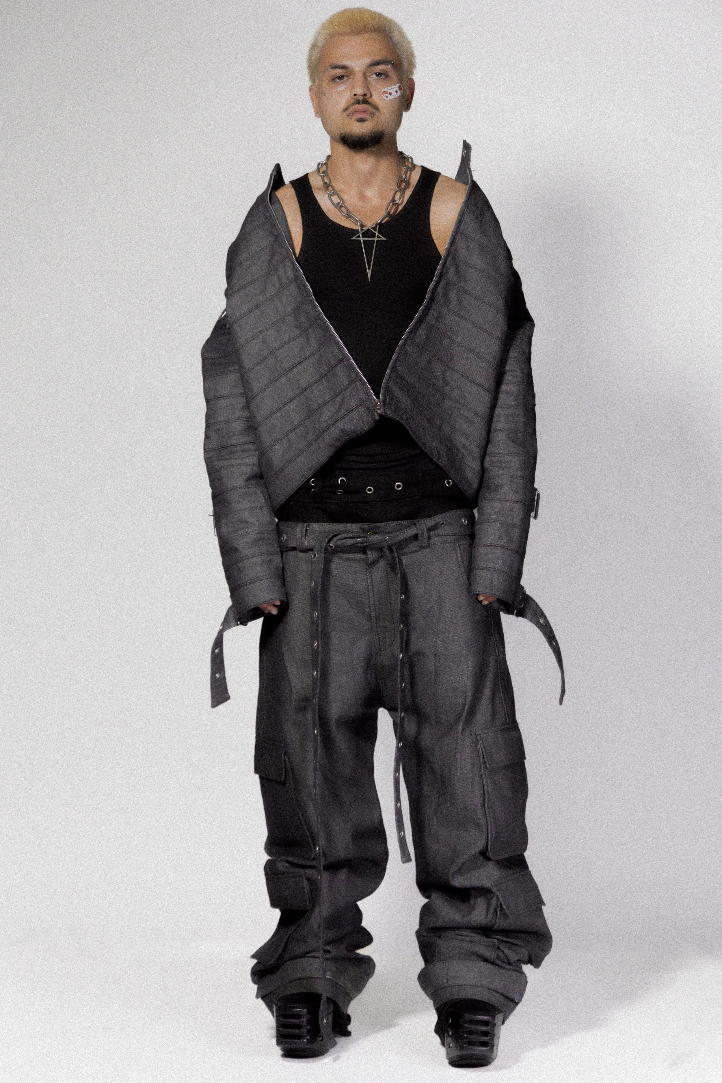 A person stands facing forward, wearing a black sleeveless shirt, oversized gray cargo pants, a gray puffer jacket draped off their shoulders, black platform shoes, and layered chains around their neck, with platinum blonde hair and a facial sticker