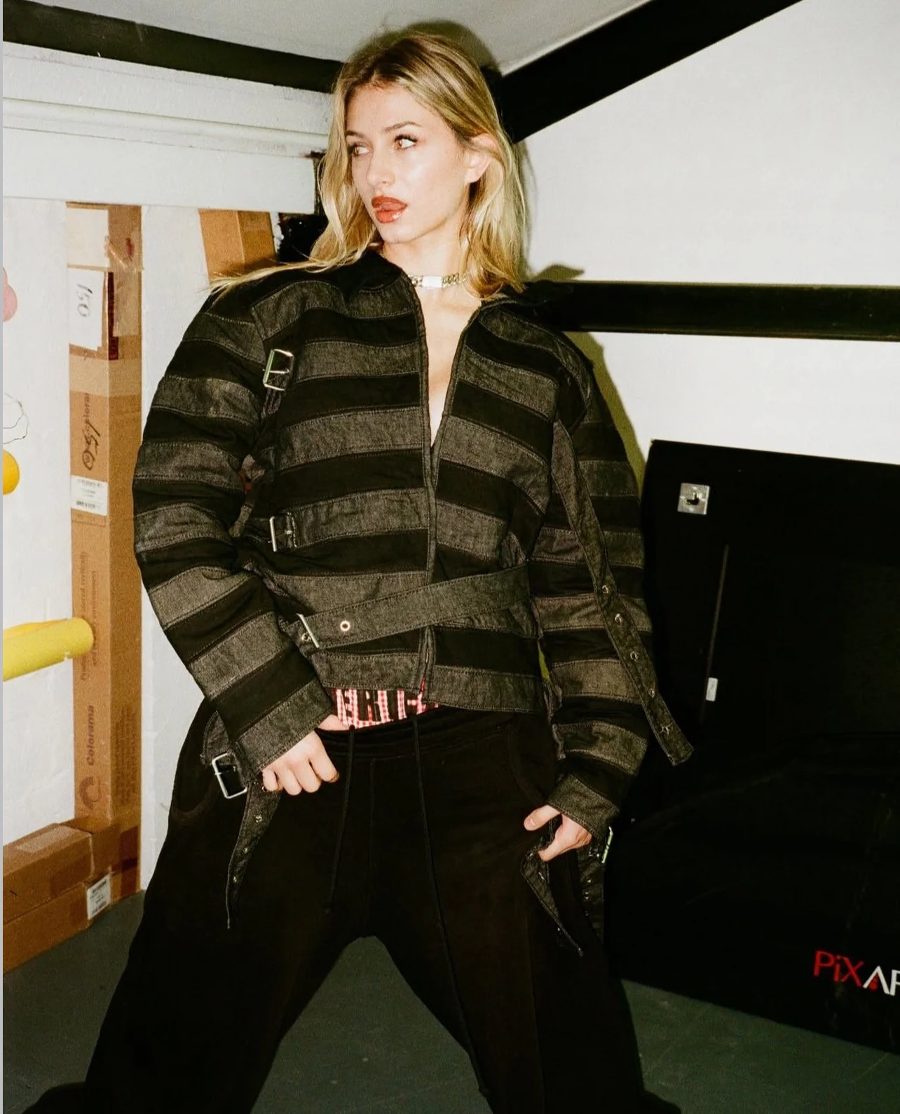 A woman wearing a black and gray striped jacket, black pants, and a choker necklace, standing in a room with cardboard boxes and a black equipment case.