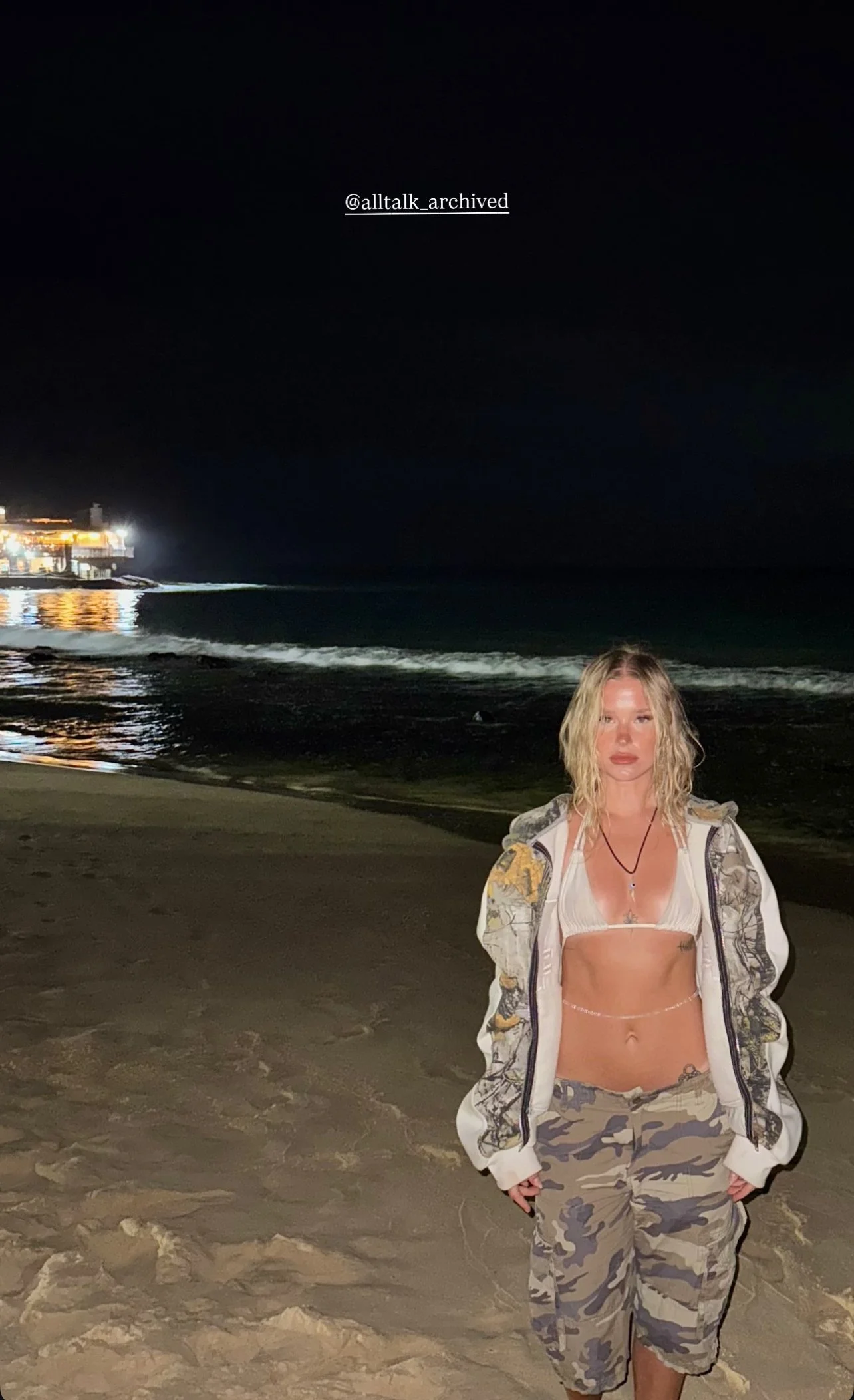 Nighttime beach scene with a woman standing on the sand in camouflage pants and a bikini top, with the ocean and a lit pier in the background.