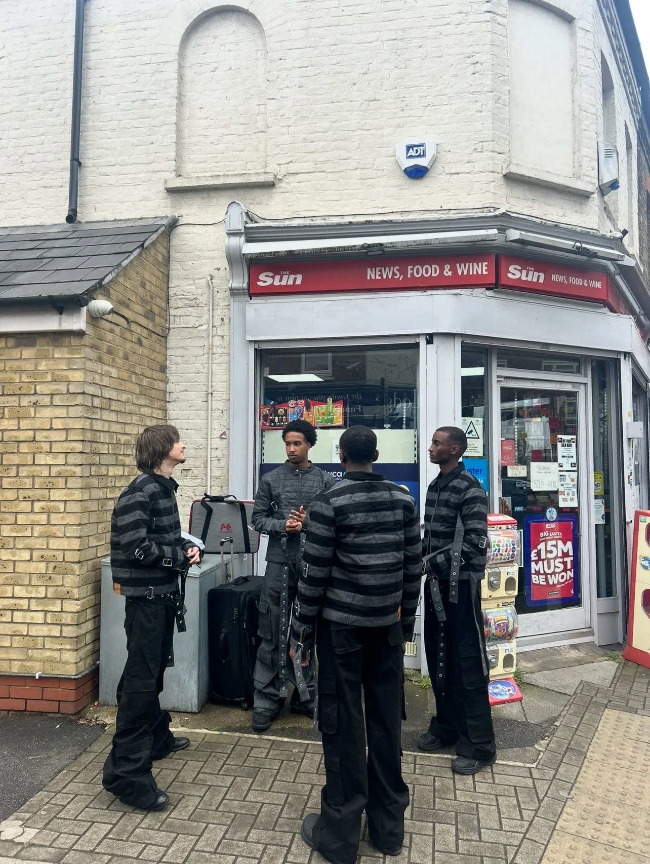 Four young men dressed in black and gray clothing talking outside a convenience store named 'The Sun' with a sign for news, food, and wine, on a city sidewalk.