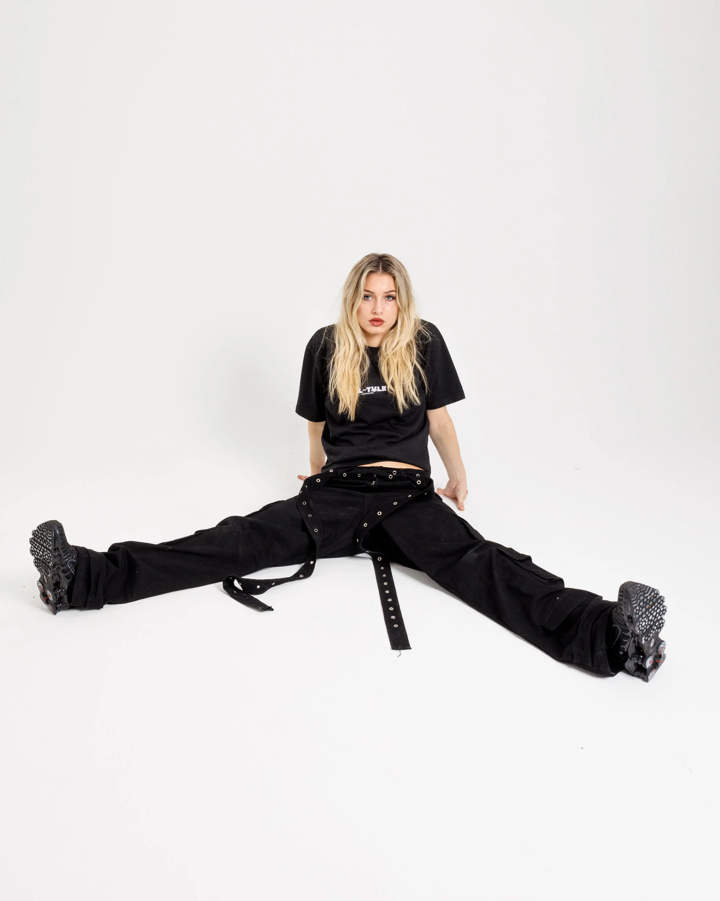 A young woman dressed in black, sitting on the floor with her legs spread apart and leaning back on her hands. She has blonde hair and is looking directly at the camera with a serious expression. The background is plain white.
