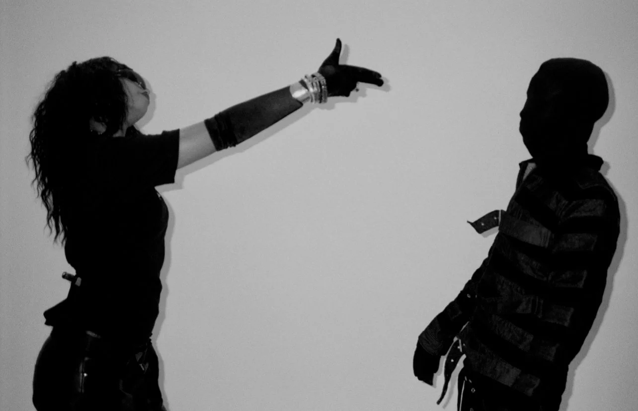 A woman with curly hair, wearing multiple bangles, points at a young boy's face with her arm extended. The boy is wearing a striped jacket. Their silhouettes are visible against a plain background.