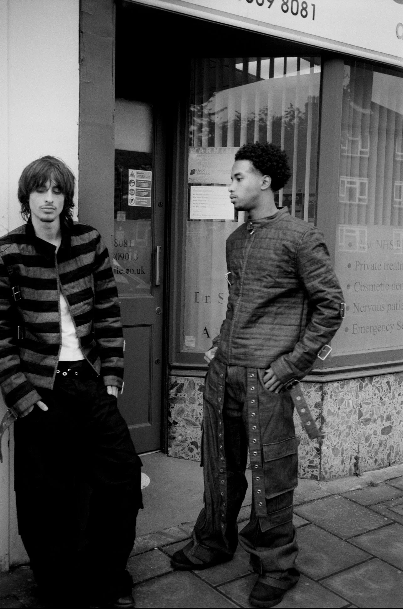 Two young men stand outside a storefront, one with long hair wearing a striped jacket and the other with curly hair in a dark jacket and baggy pants, both with casual poses.