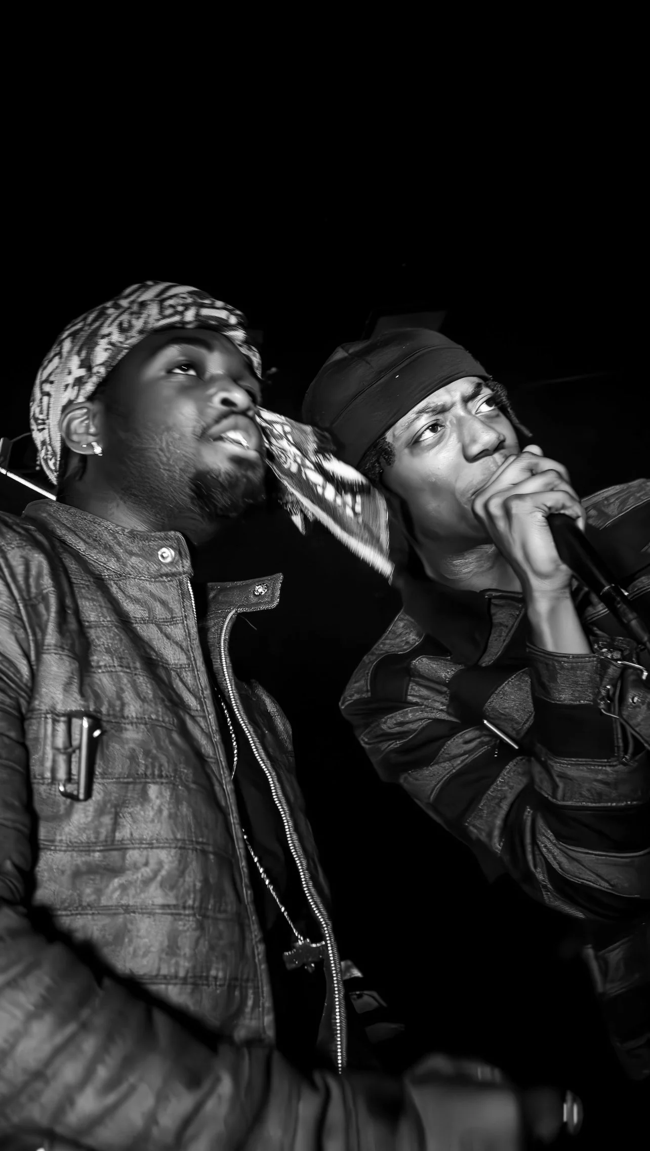 Two men appear to be performing together on stage, with one holding a microphone. They are dressed in stylish jackets and head coverings, and are captured in black and white during a night event.