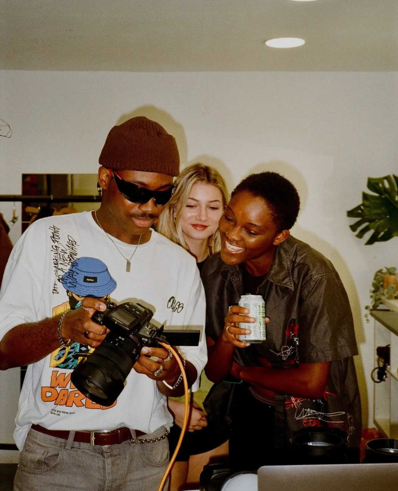 Three women gather around a camera, looking at the screen, smiling, with one holding a drink, in a casual indoor setting.