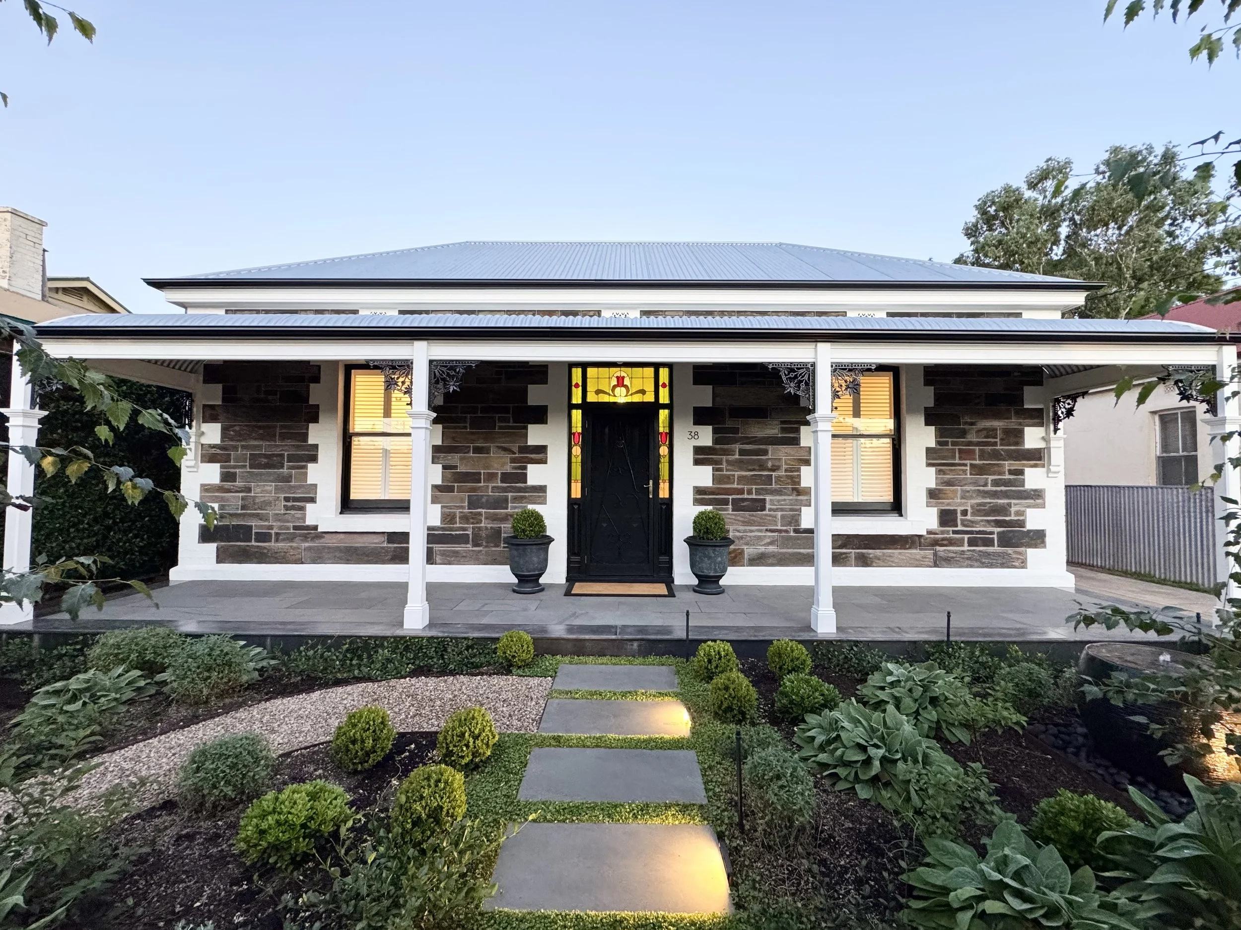 Front view of a house with a stone facade, white trim, black door with stained glass, and a covered porch. There are potted plants, landscaped garden with stepping stones, and some trees around the house.