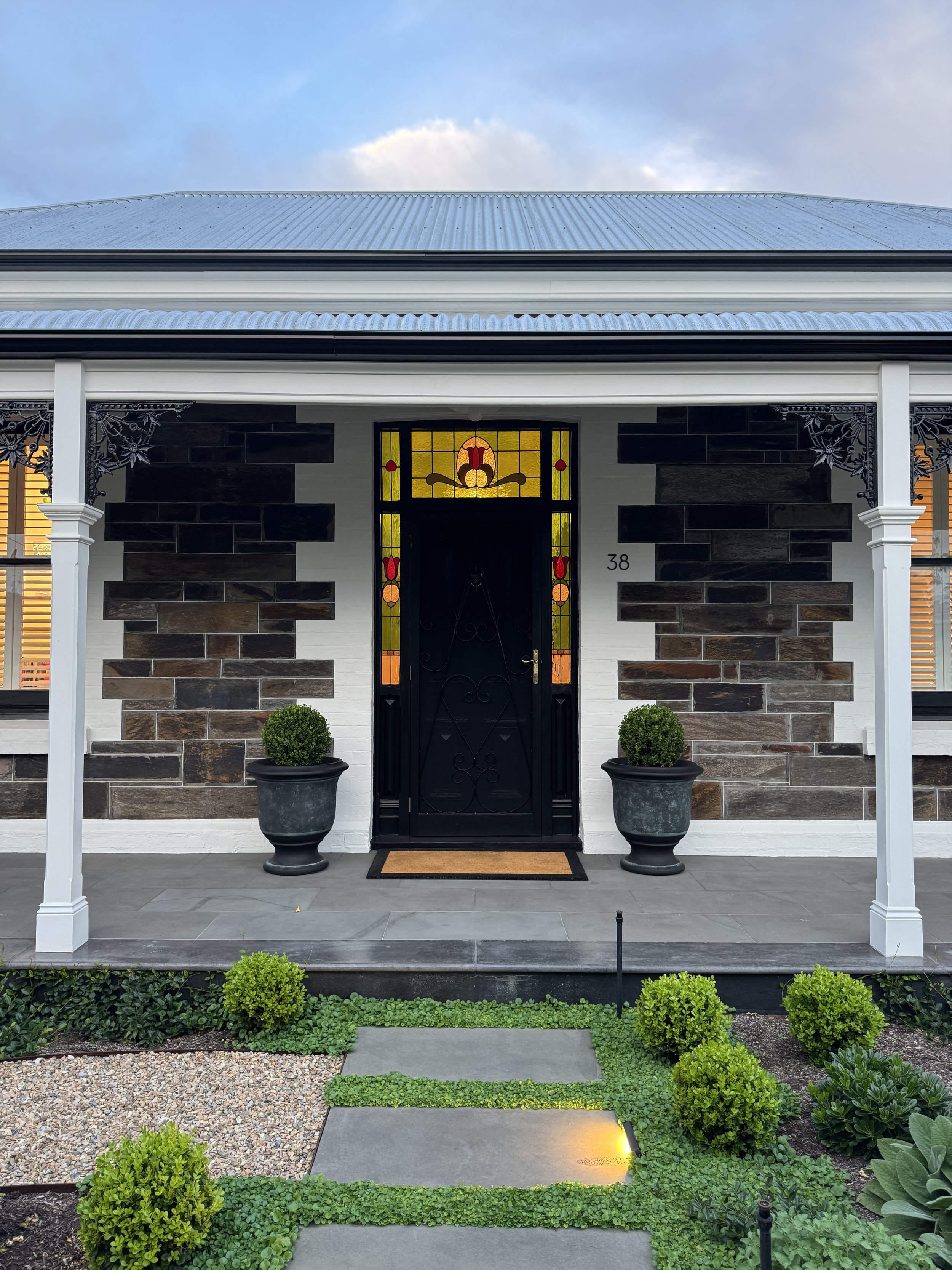 Front view of a house with a black door featuring stained glass, two potted bushes by the doorway, a small garden with green shrubs, and a stone pathway leading to the entrance.