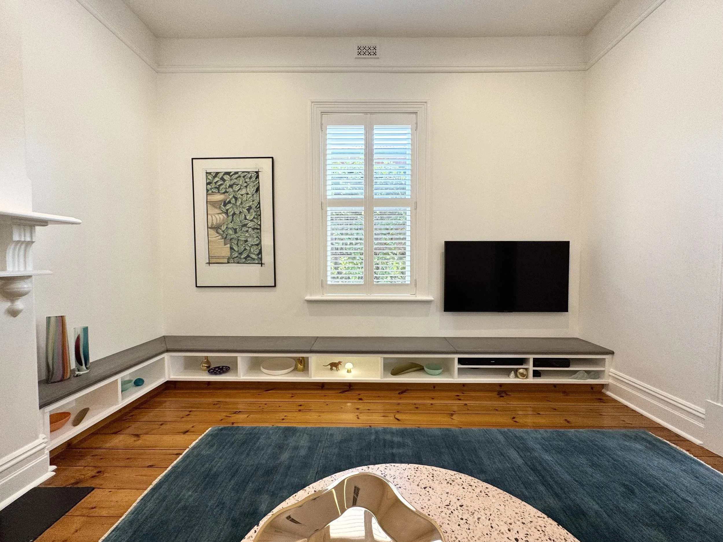 Living room with white walls, a window with shutters, a wall-mounted TV, a framed artwork, a built-in shelf with decorative objects, and a blue rug.