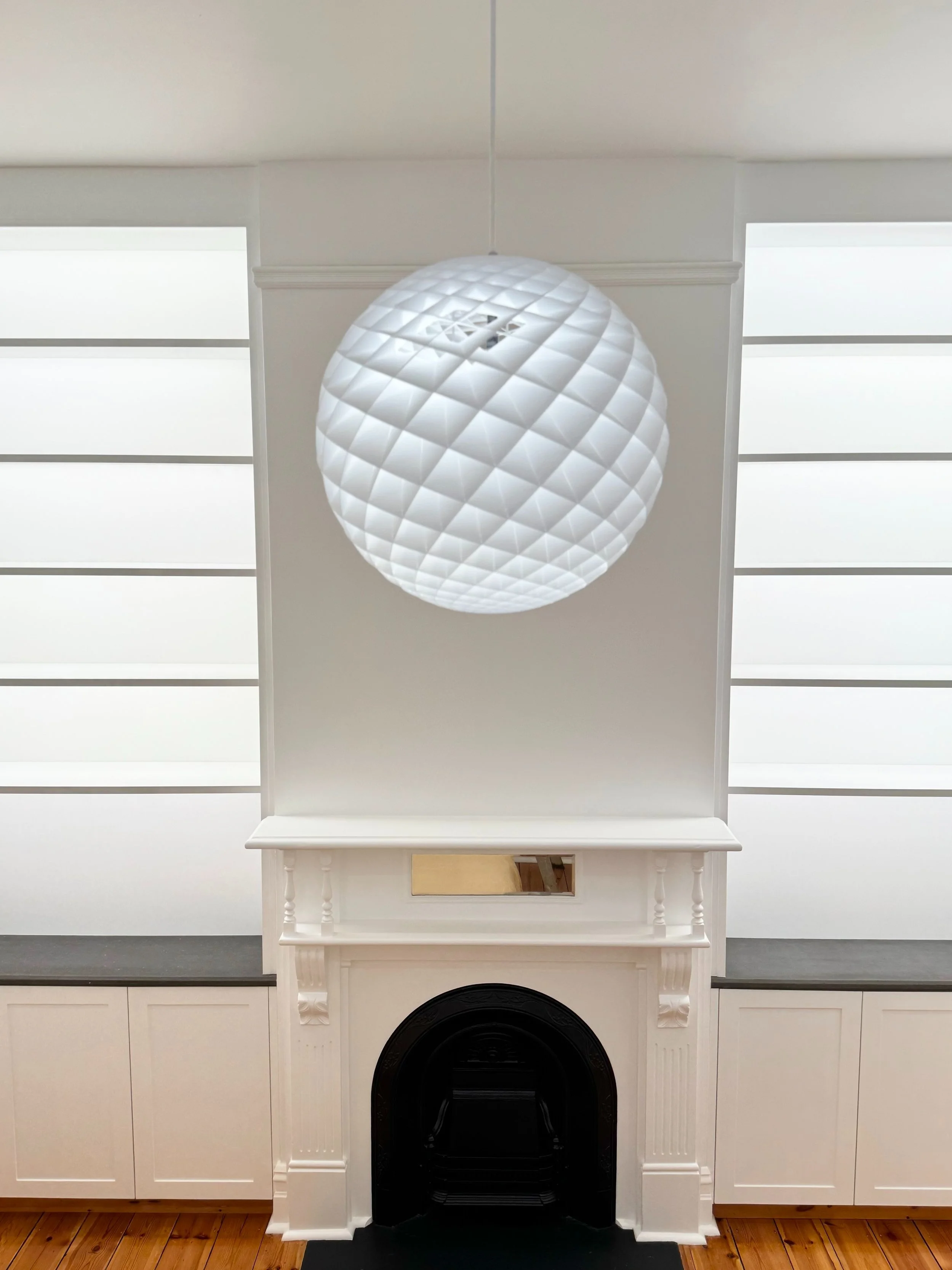 Interior of a room with a white fireplace, a mirror above it, black hearth, wooden floor, and a white geometric paper lantern hanging from the ceiling.