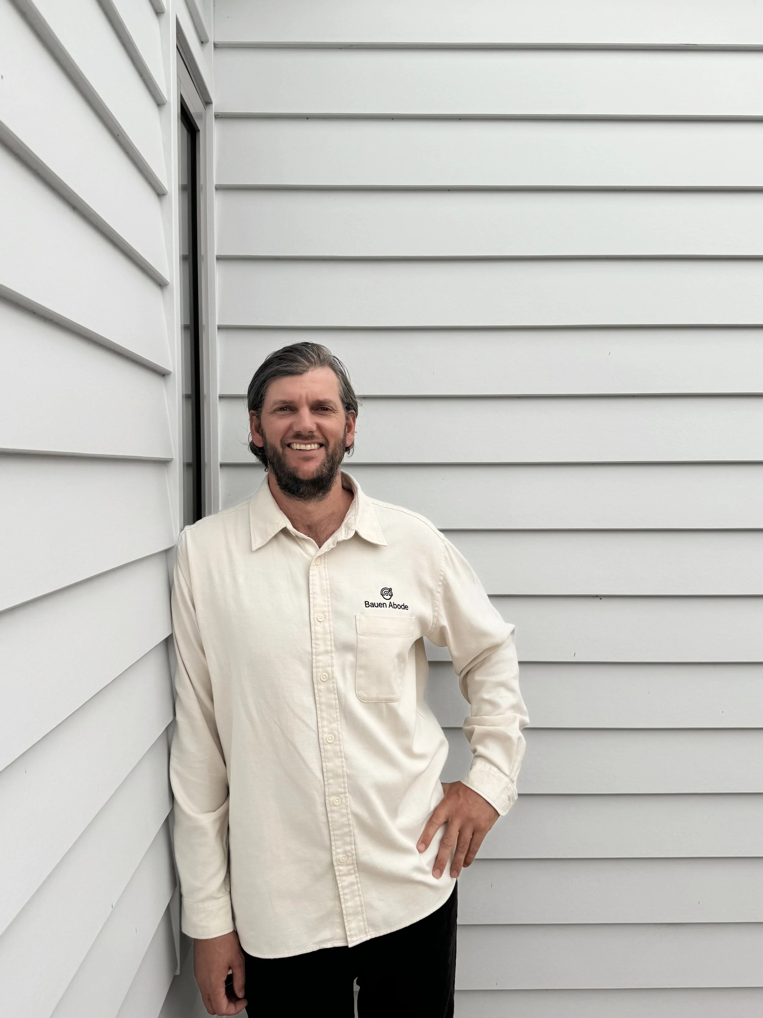 A man with dark hair and a beard, wearing a cream-colored shirt with a logo on the pocket, stands smiling against the corner of two white house walls with horizontal siding.