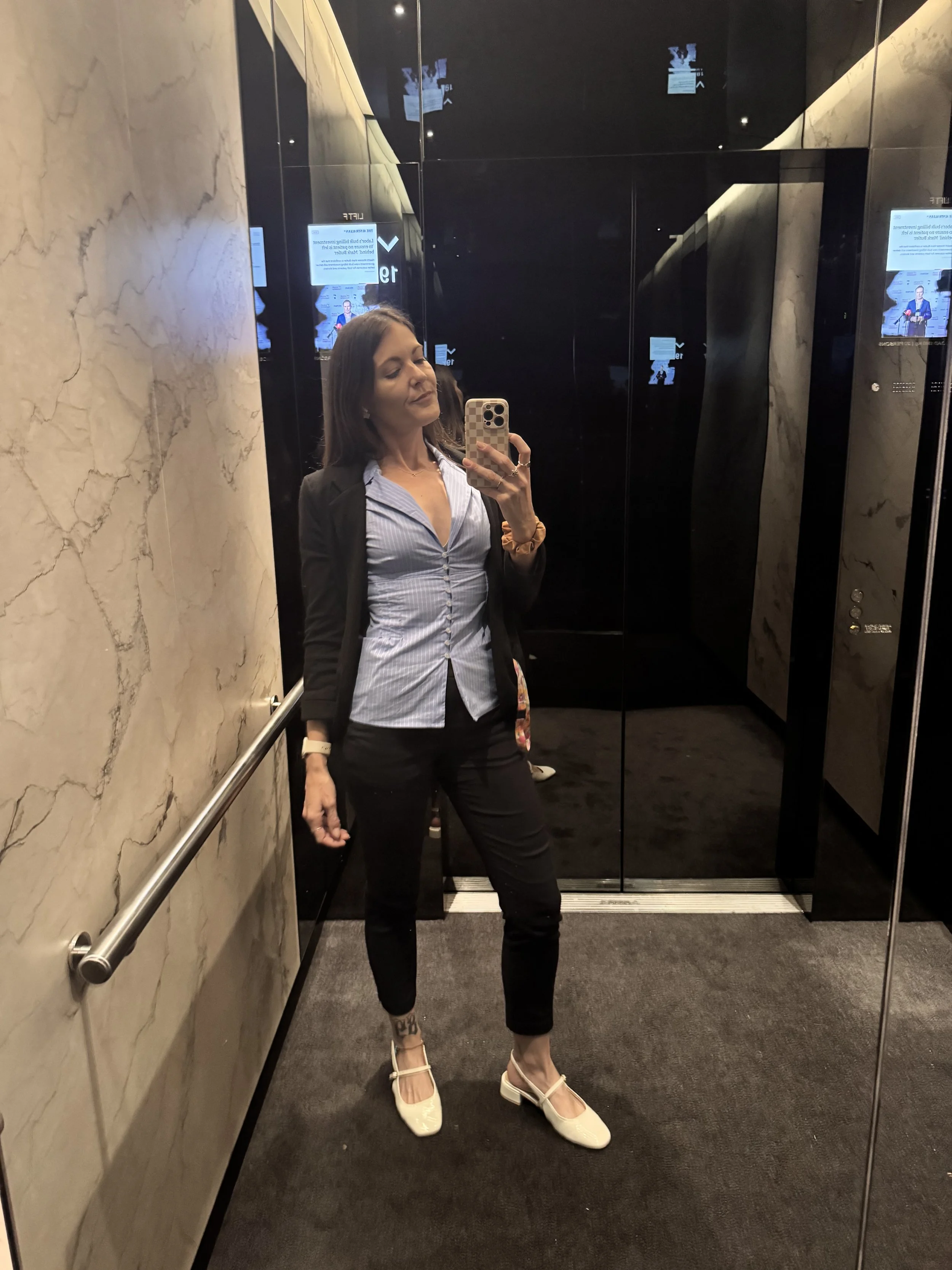 A woman taking a mirror selfie in an elevator. She is wearing a black blazer, a light blue striped blouse, black pants, and cream-colored heels. She has dark hair and is holding her phone with her right hand, with reflections visible on the elevator'