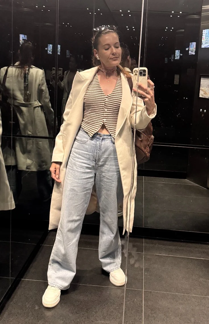 Woman taking a mirror selfie in a fitting room, wearing a beige coat, patterned crop top, light-wash jeans, and white sneakers.