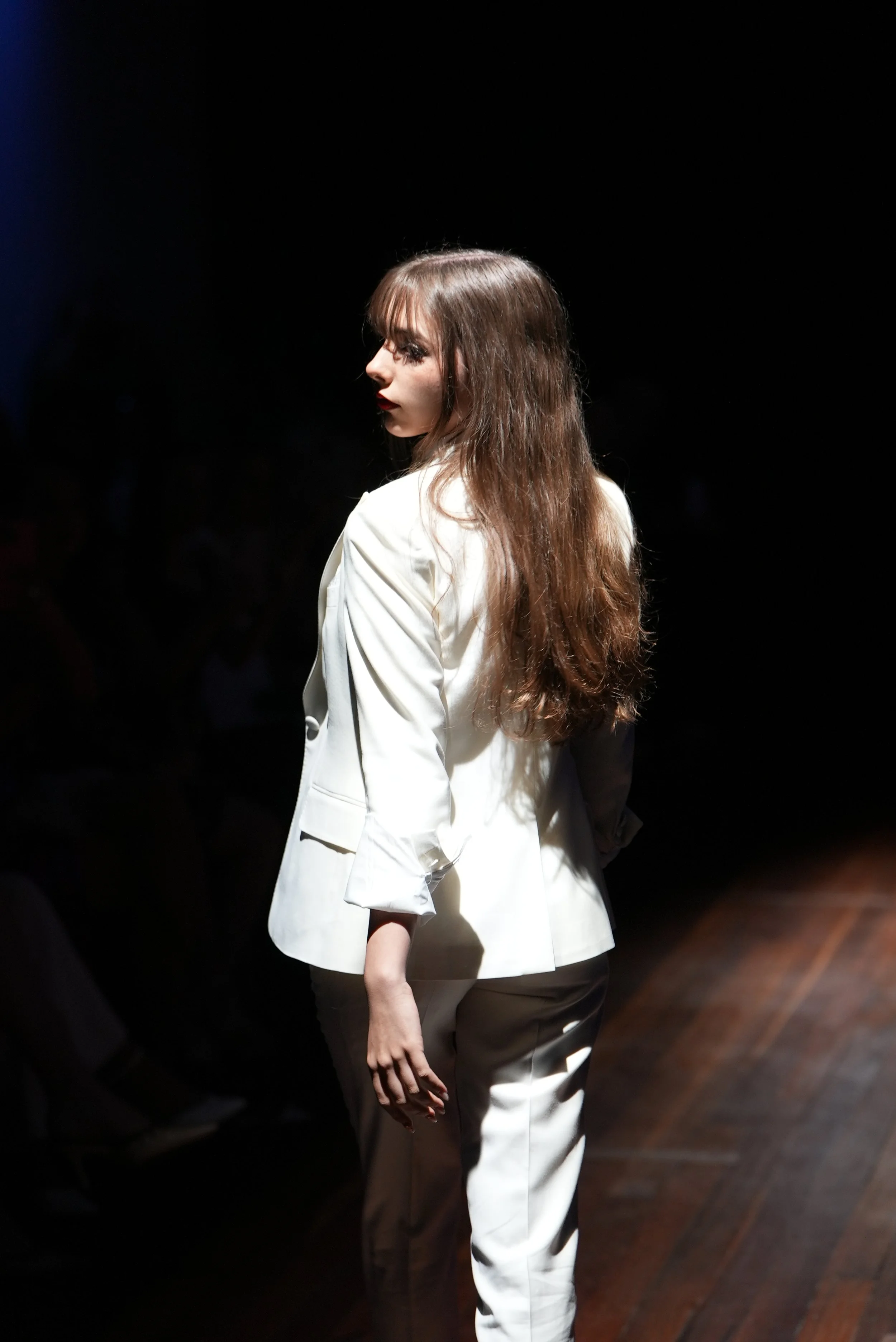 A woman walking on a runway during a fashion show, illuminated by bright lights, with a dark background.