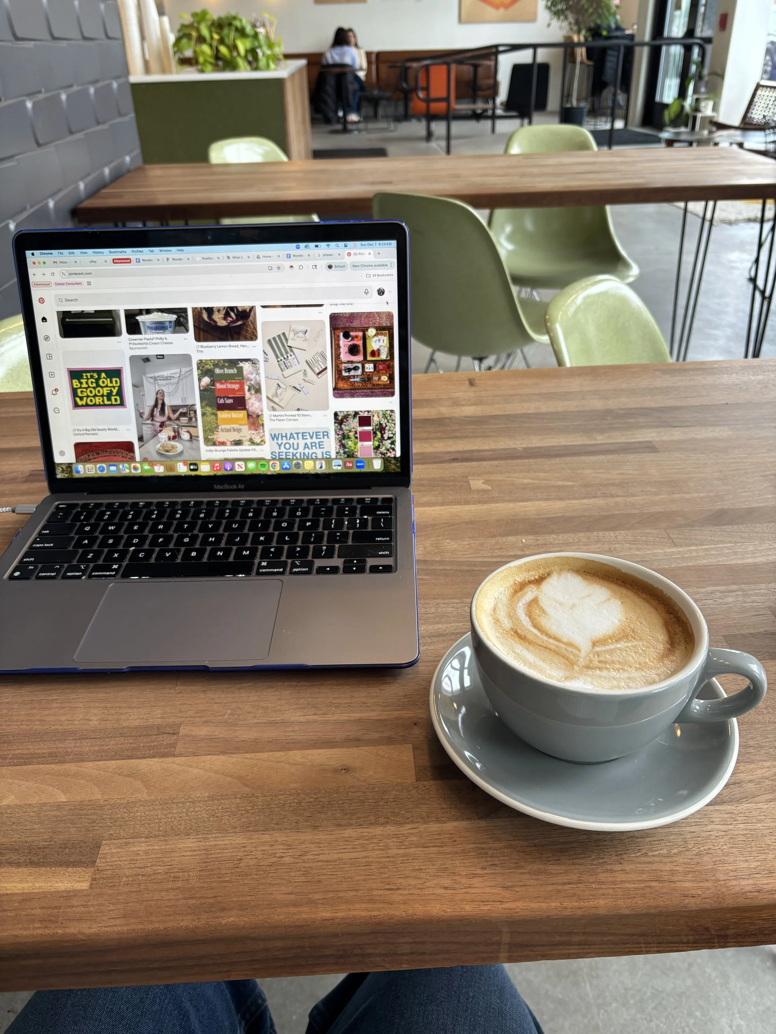 A wooden table with a laptop displaying Pinterest, a cup of latte with foam art, in a cozy cafe with green chairs, large windows, and a person working in the background.