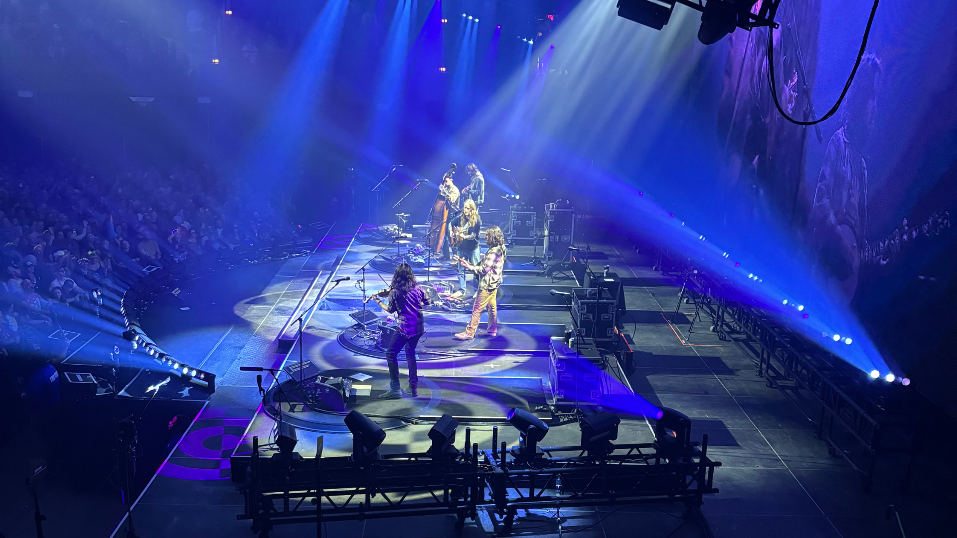 A band performs on a stage with blue and purple lighting in front of a large audience.