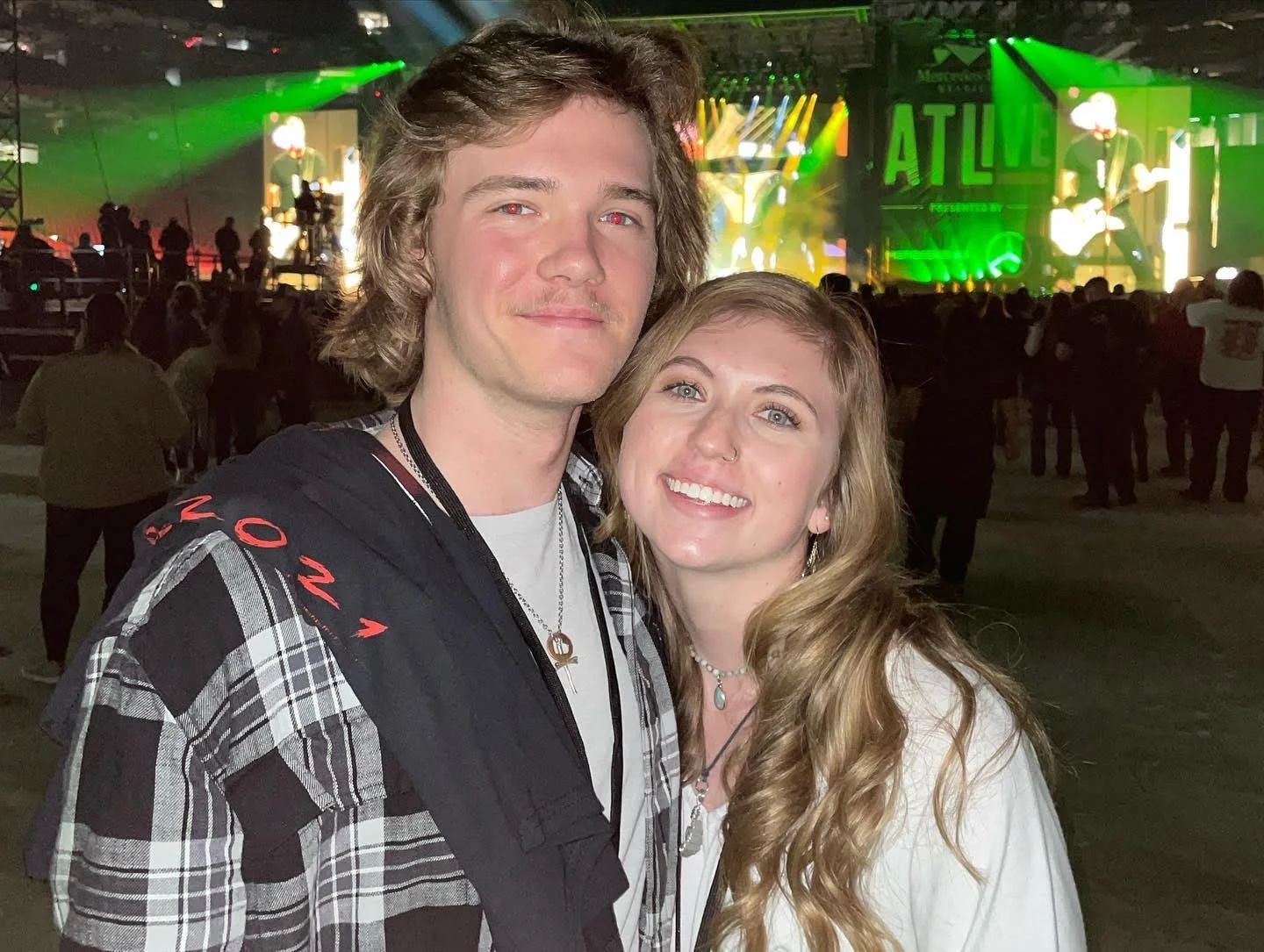 A young man and woman smiling at the camera at a concert or music festival, with a stage featuring bright green lighting and a crowd in the background.