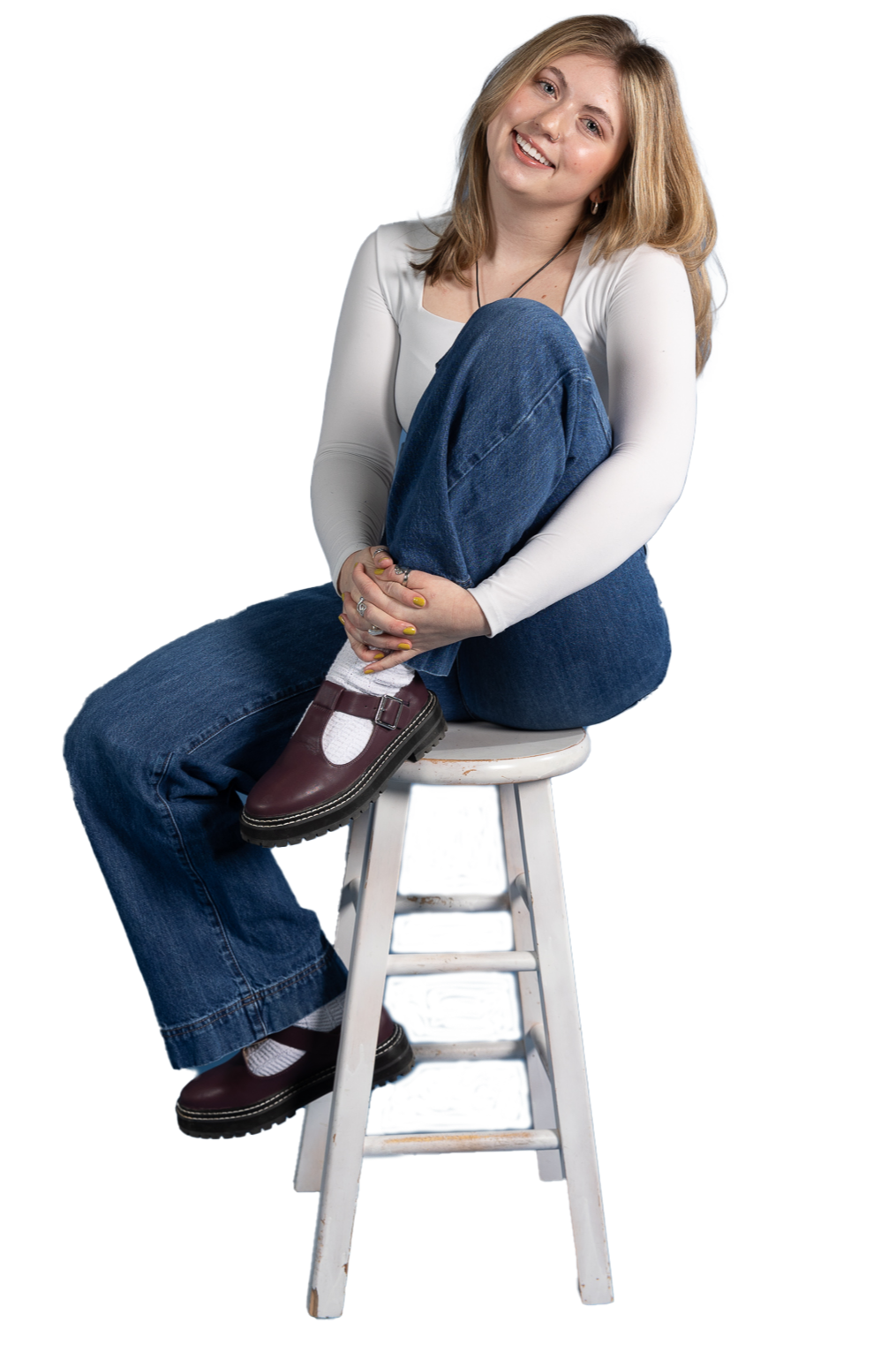 A young woman with blonde hair sits on a white stool, smiling at the camera. She is dressed in a white long-sleeve shirt, blue jeans, and burgundy shoes with white socks.